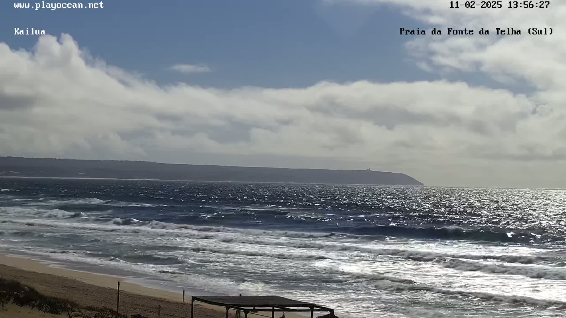 A partly cloudy day shows a sandy beach with moderate waves rolling onto the shore, and a distant landmass visible across the ocean.