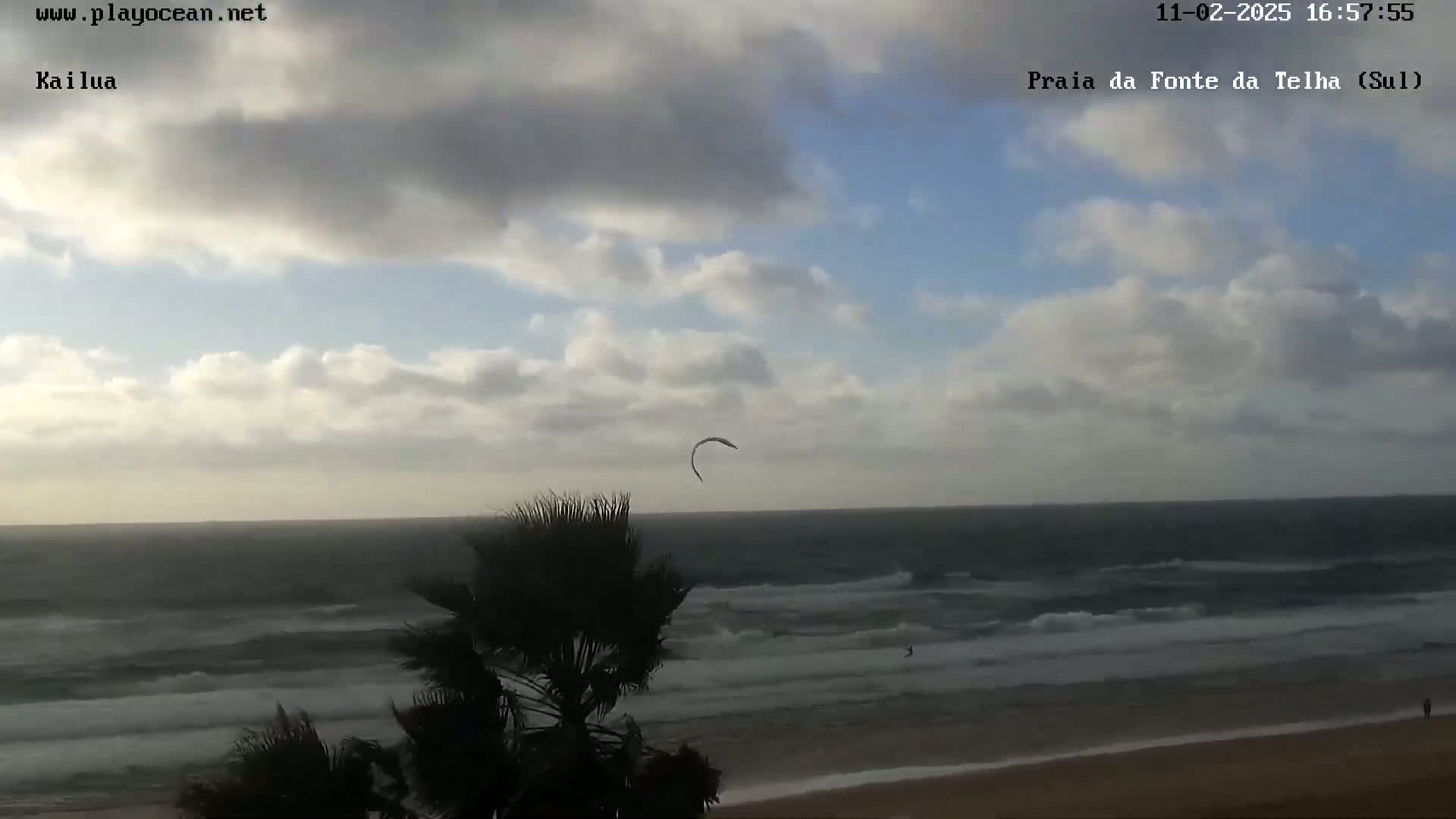 A kite surfer is visible in the distance over a wavy ocean under a partly cloudy sky.