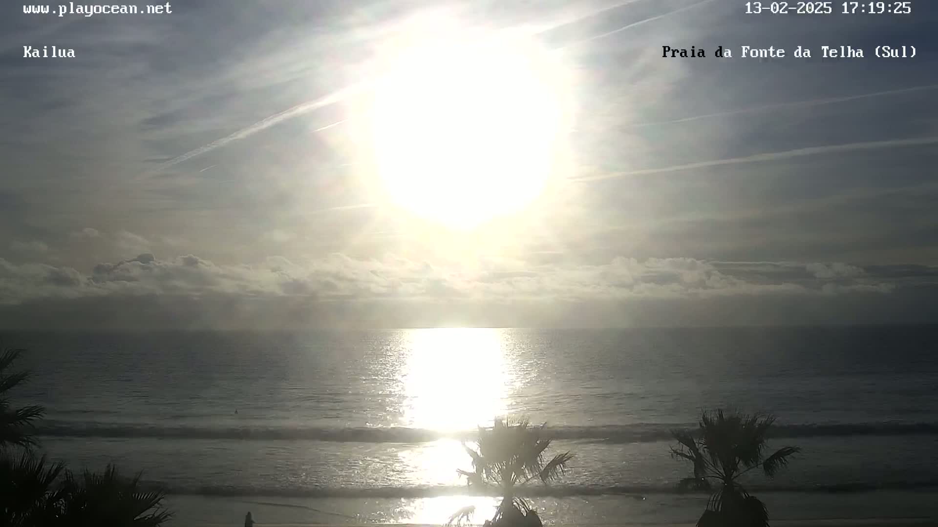A bright sun shines over a calm ocean with a few clouds in the sky, partially obscured by palm trees on the beach.
