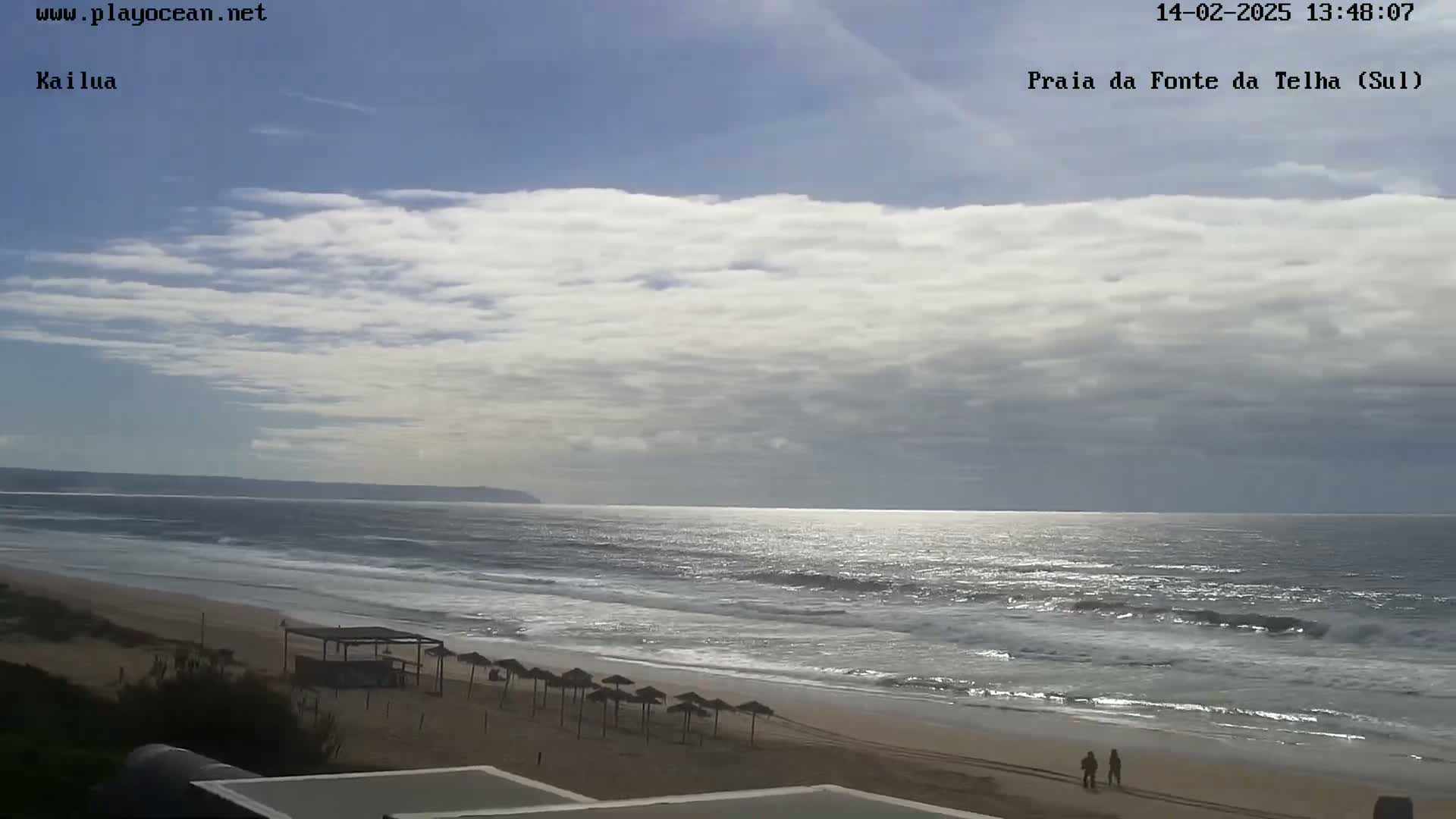 A partly cloudy day shows an ocean beach with some small waves, a few beach umbrellas, and two people walking near the water's edge.