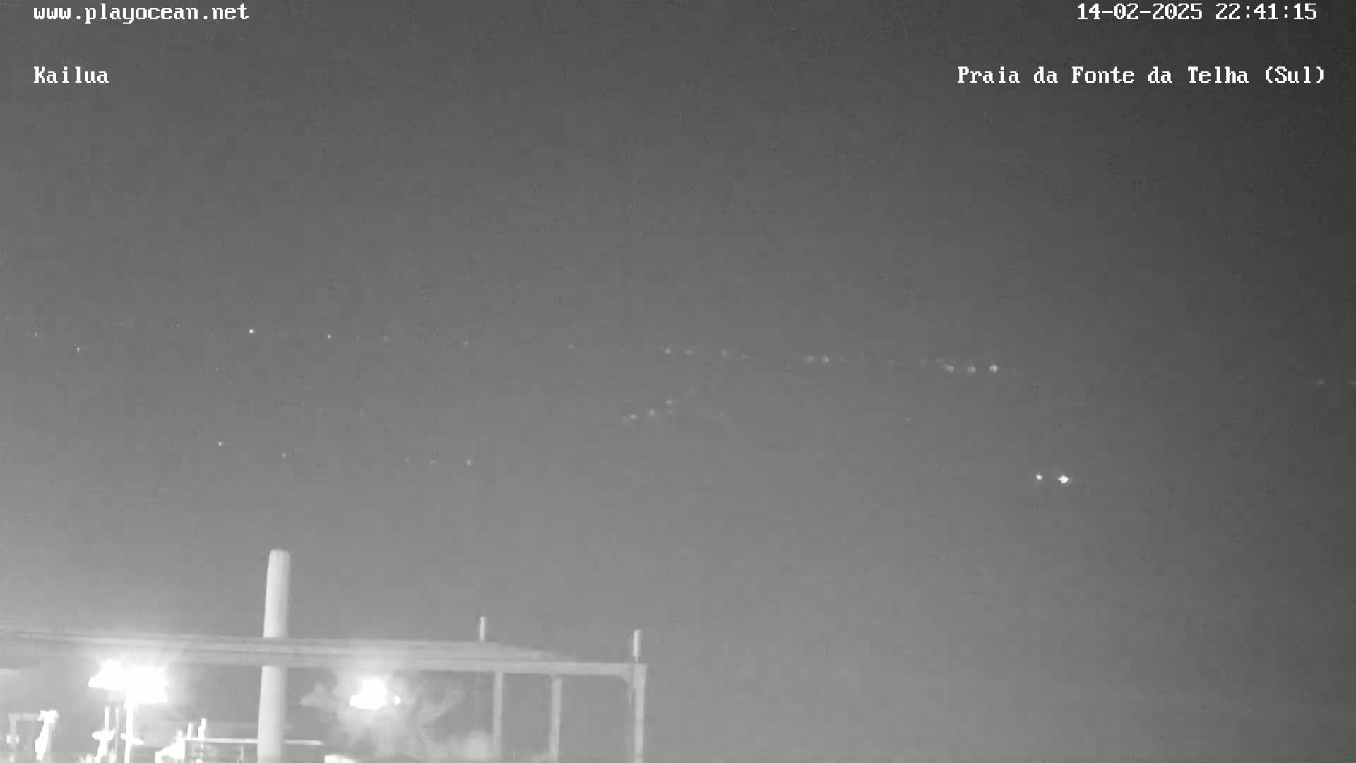 A nighttime, outdoor view shows a line of lights in the distance above a dark structure with people near lights on it.