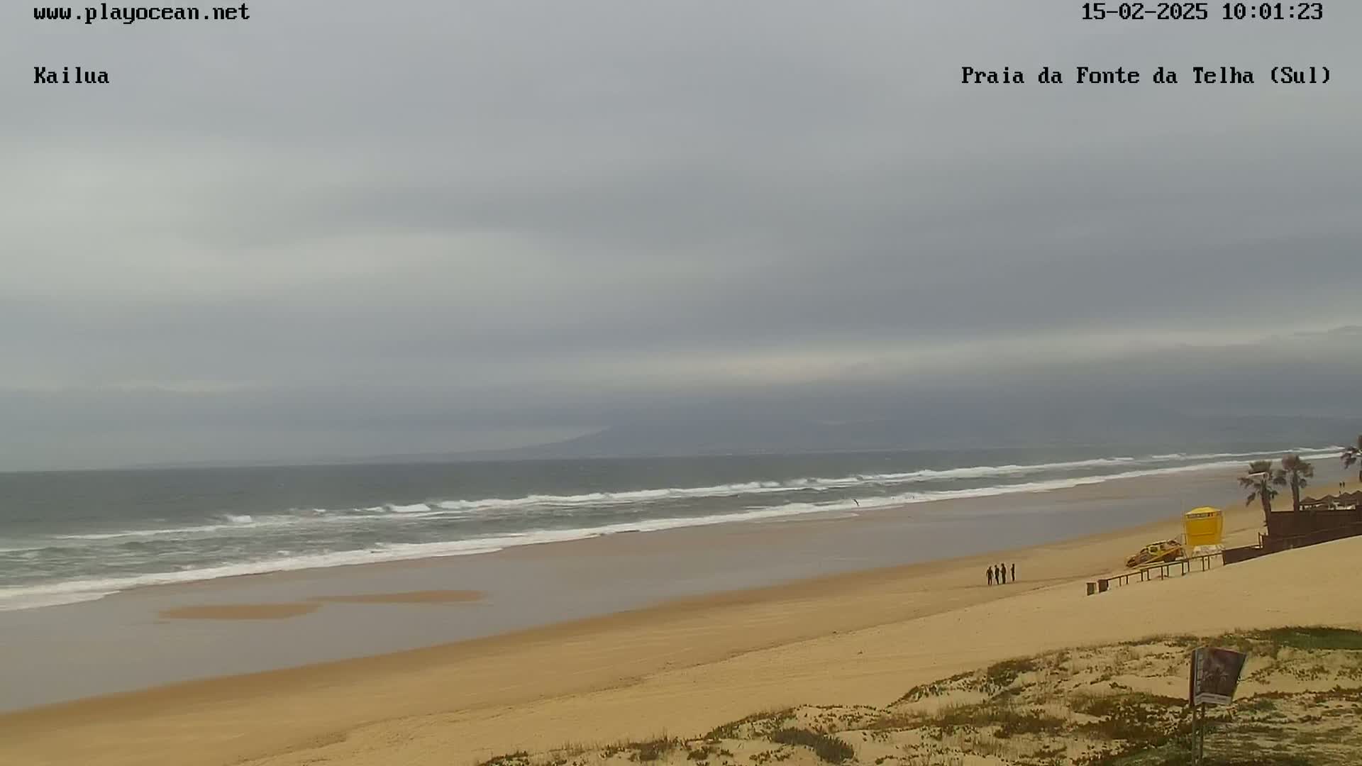 A sandy beach with a calm ocean under an overcast sky shows a few people walking near a lifeguard stand.