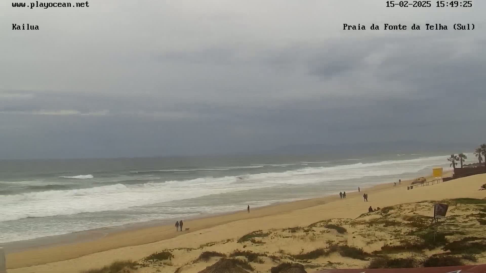 A sandy beach with a few people walking on it stretches along a gray, overcast ocean with moderate waves.