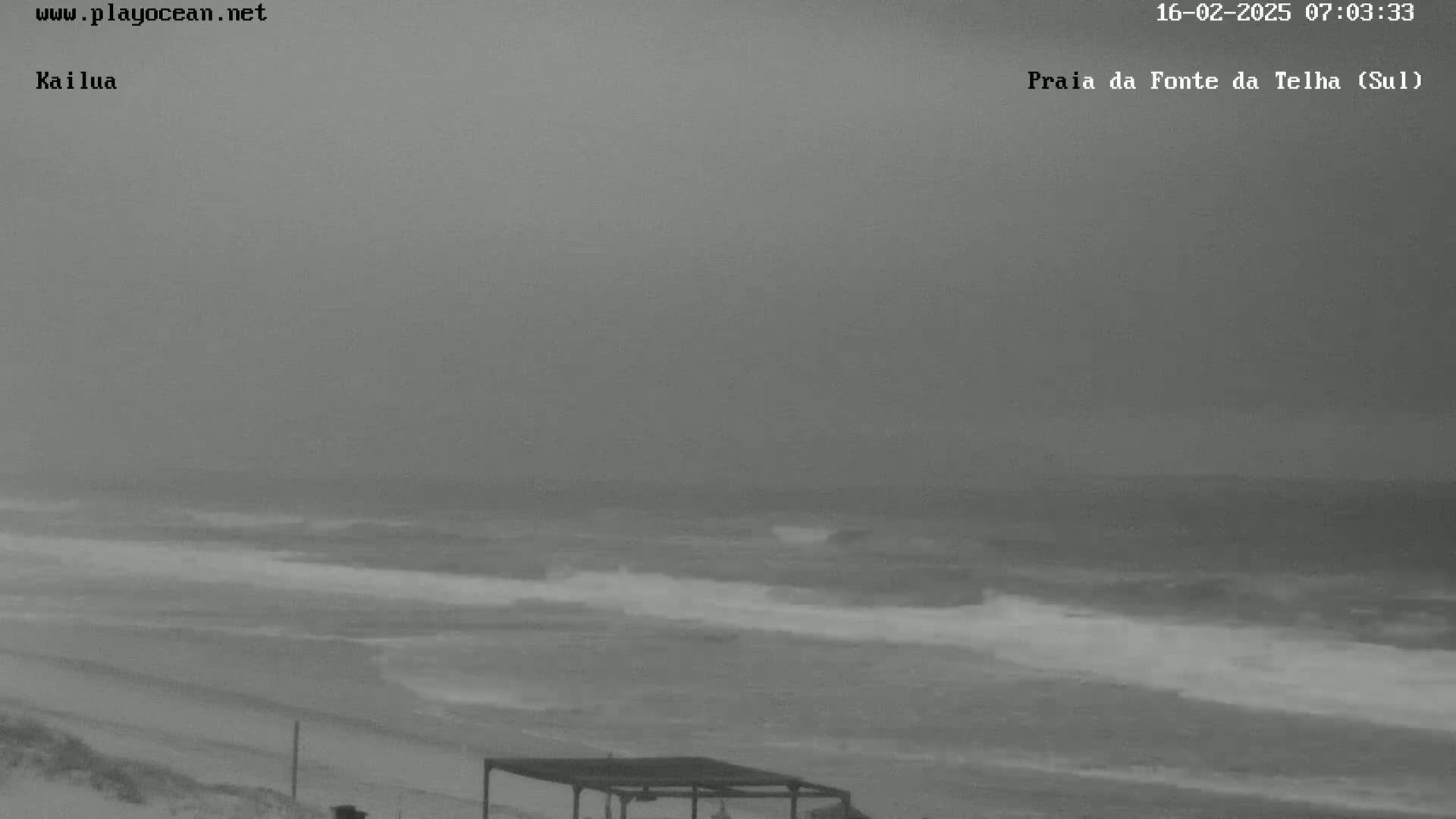 A gray, overcast day shows a beach with waves breaking on the shore, and a covered structure is visible in the foreground.