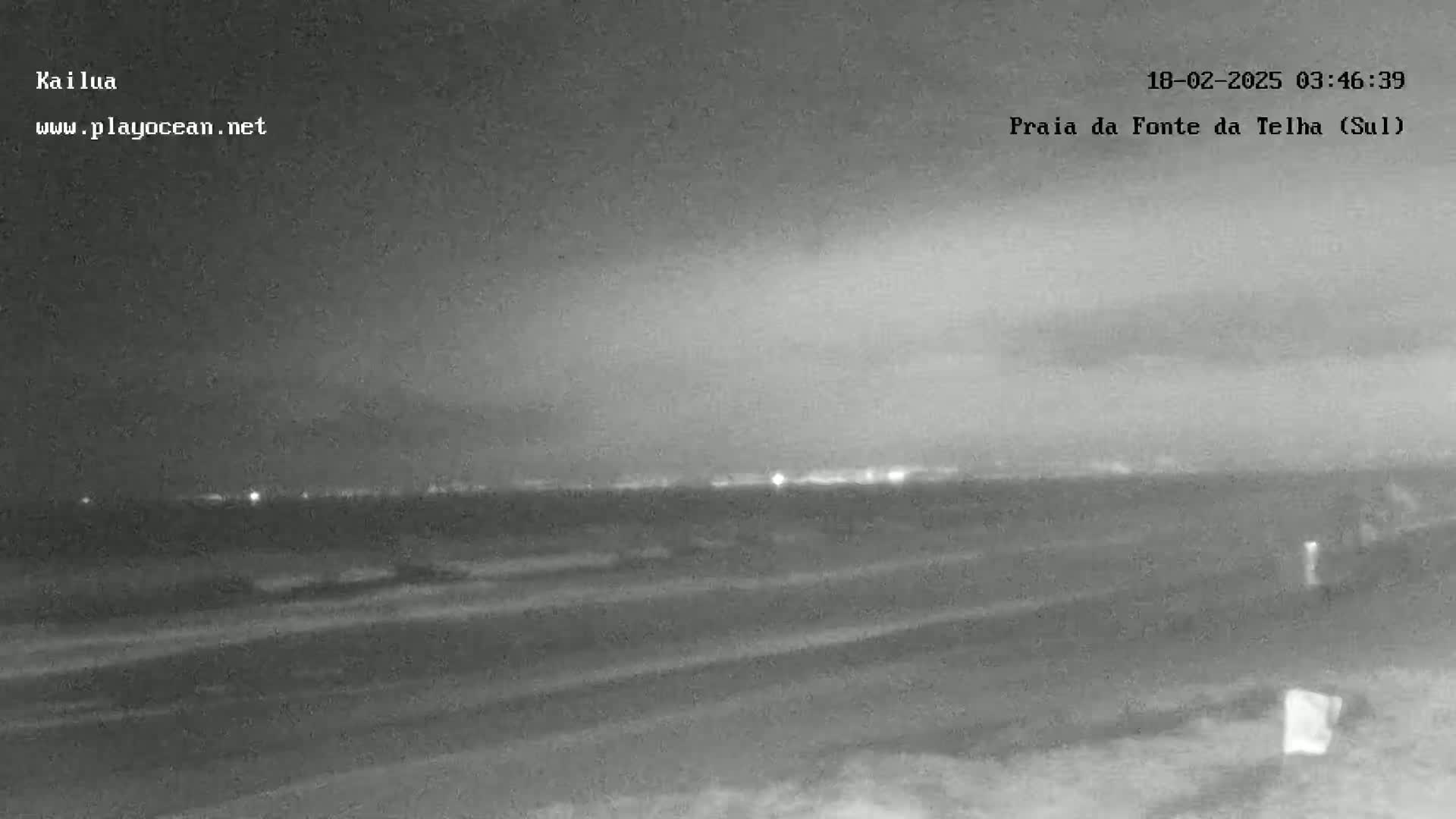 A nighttime, overcast view shows a dark beach and a distant, faintly lit horizon.