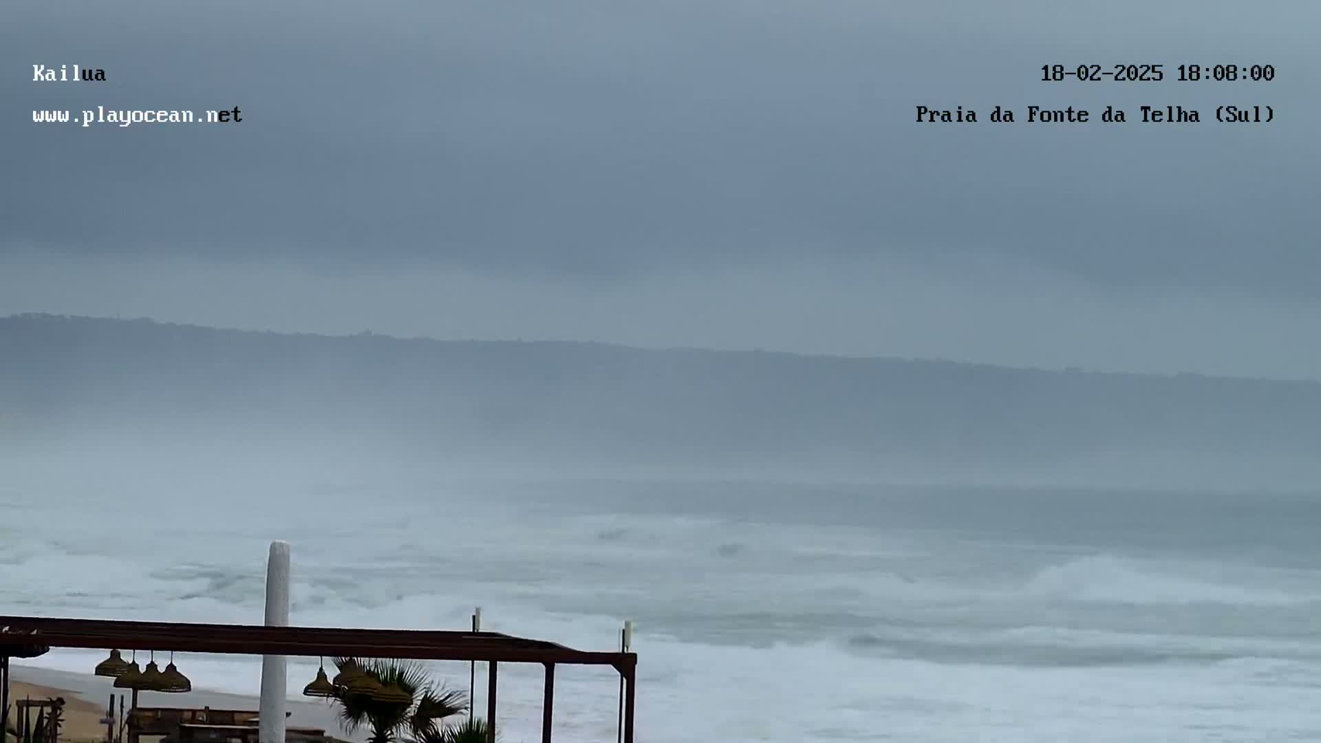 A hazy, overcast day shows a beach with somewhat choppy waves and a partially visible outdoor structure.