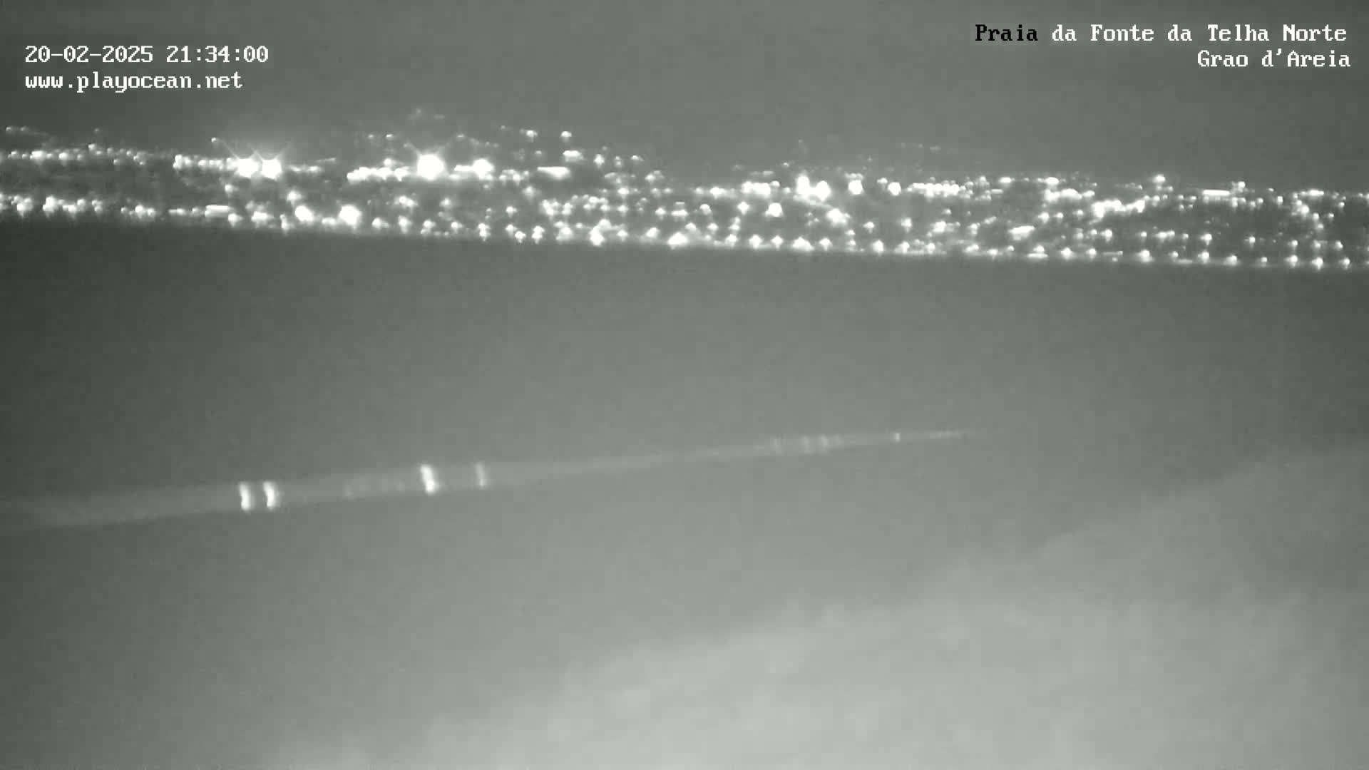 A nighttime long shot shows a distant city's lights across a dark body of water under a mostly clear sky.