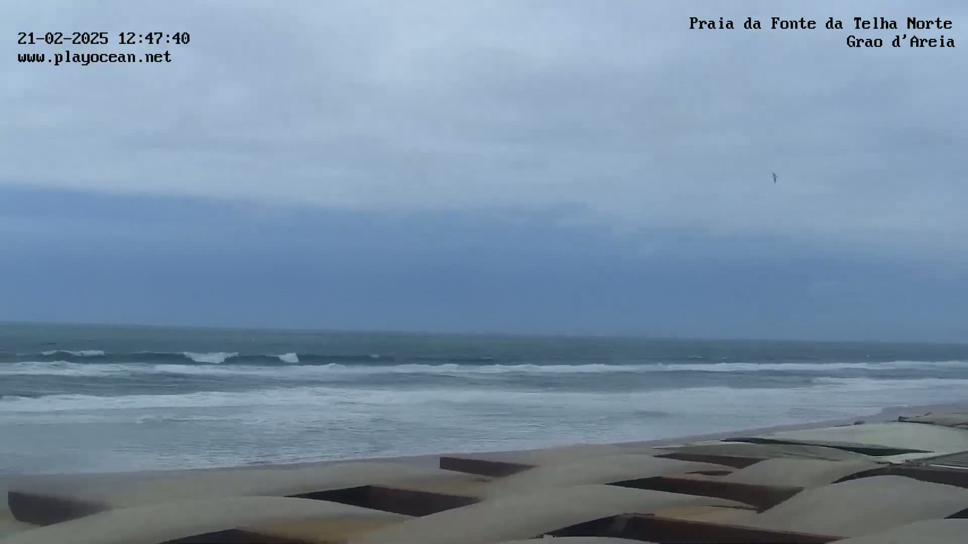 Under an overcast sky, a beach with moderate waves is partially visible behind a series of low-lying structures.