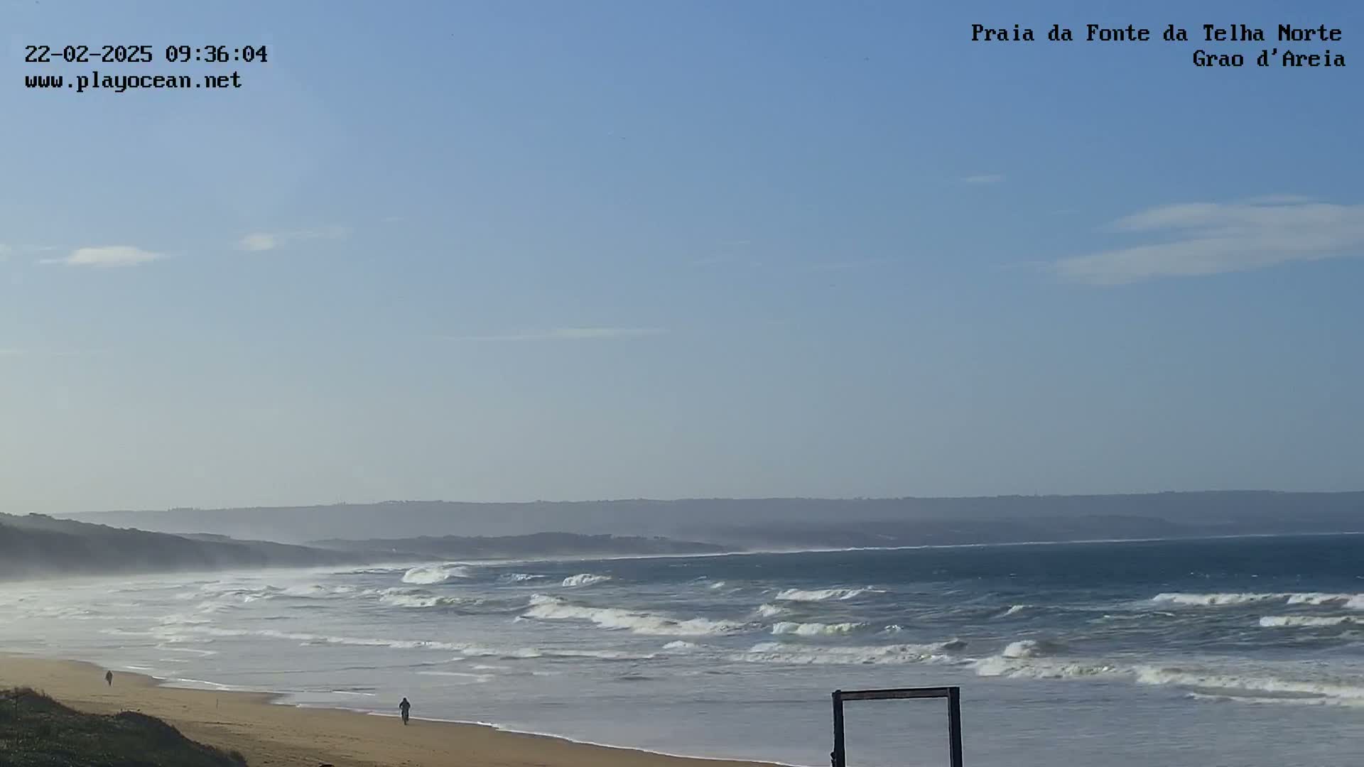 Under a mostly clear sky, a wide sandy beach stretches out to a wavy ocean with moderate surf, and a few people walk along the shore.