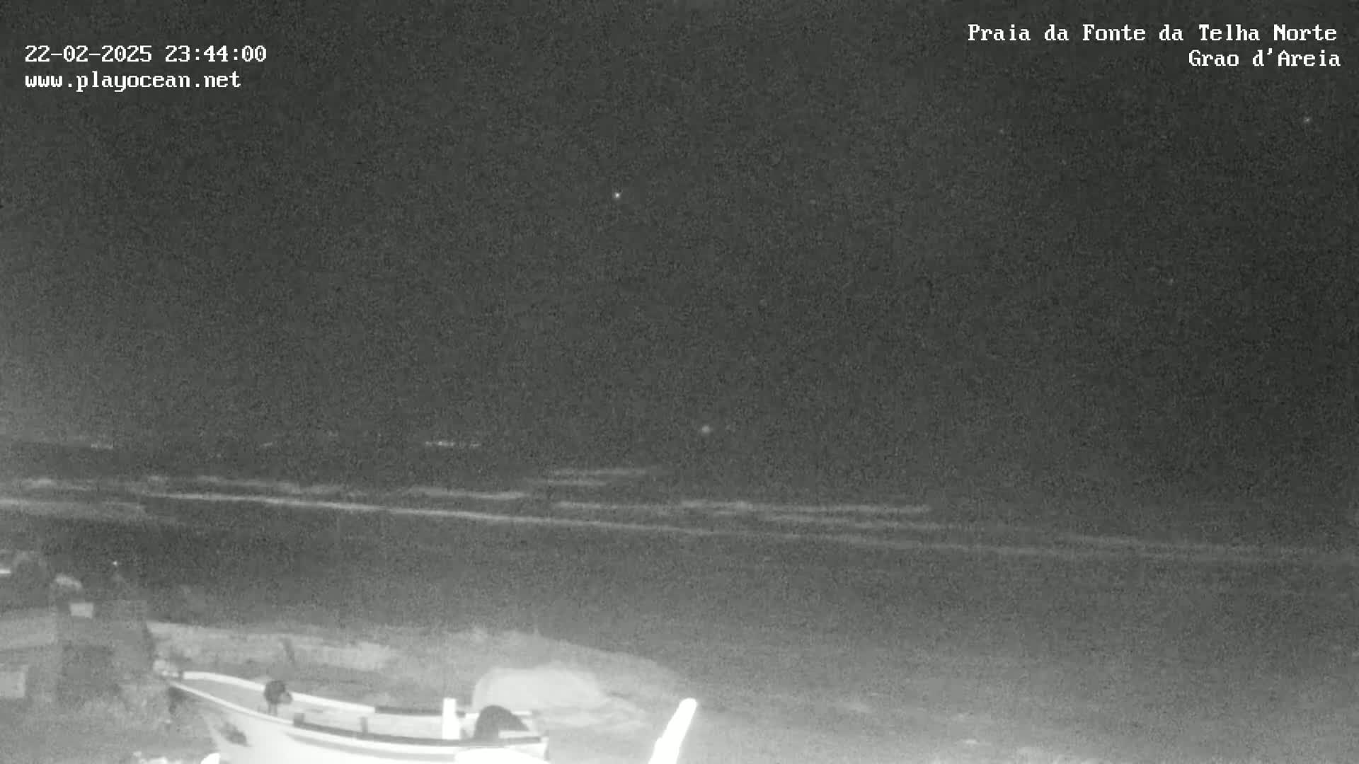A nighttime, low-light view shows a dark ocean with small waves under a clear sky, and a small boat is partially visible in the foreground on a sandy shore.