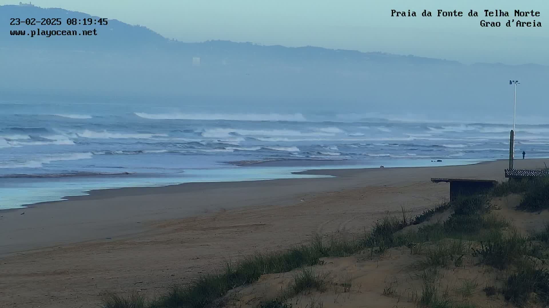 A hazy, overcast day reveals moderate waves breaking on a sandy beach backed by sand dunes and distant hills.