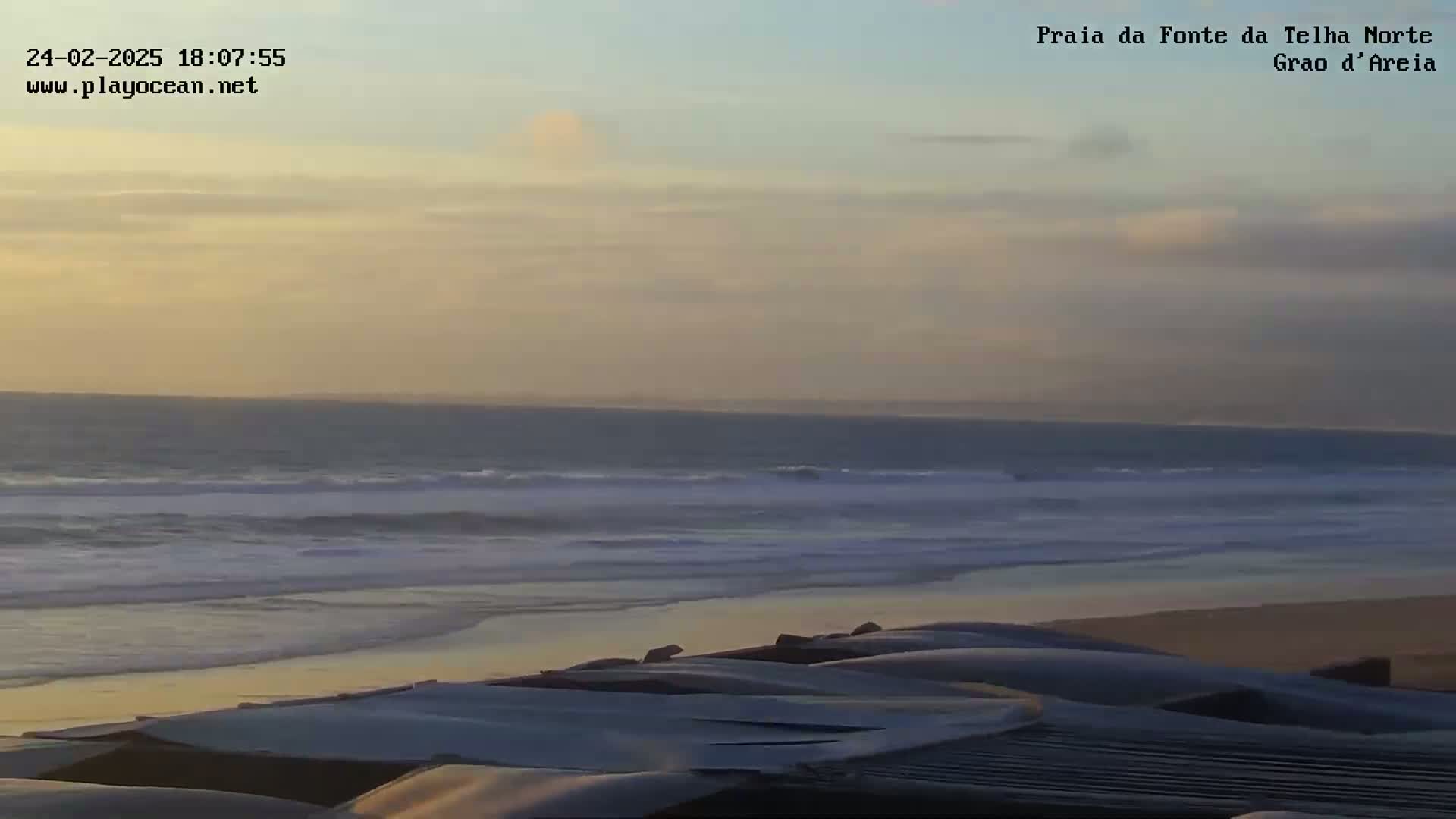 A sandy beach with calm waves under a pale sunset sky, covered with what appears to be a collection of folded fabrics or coverings.