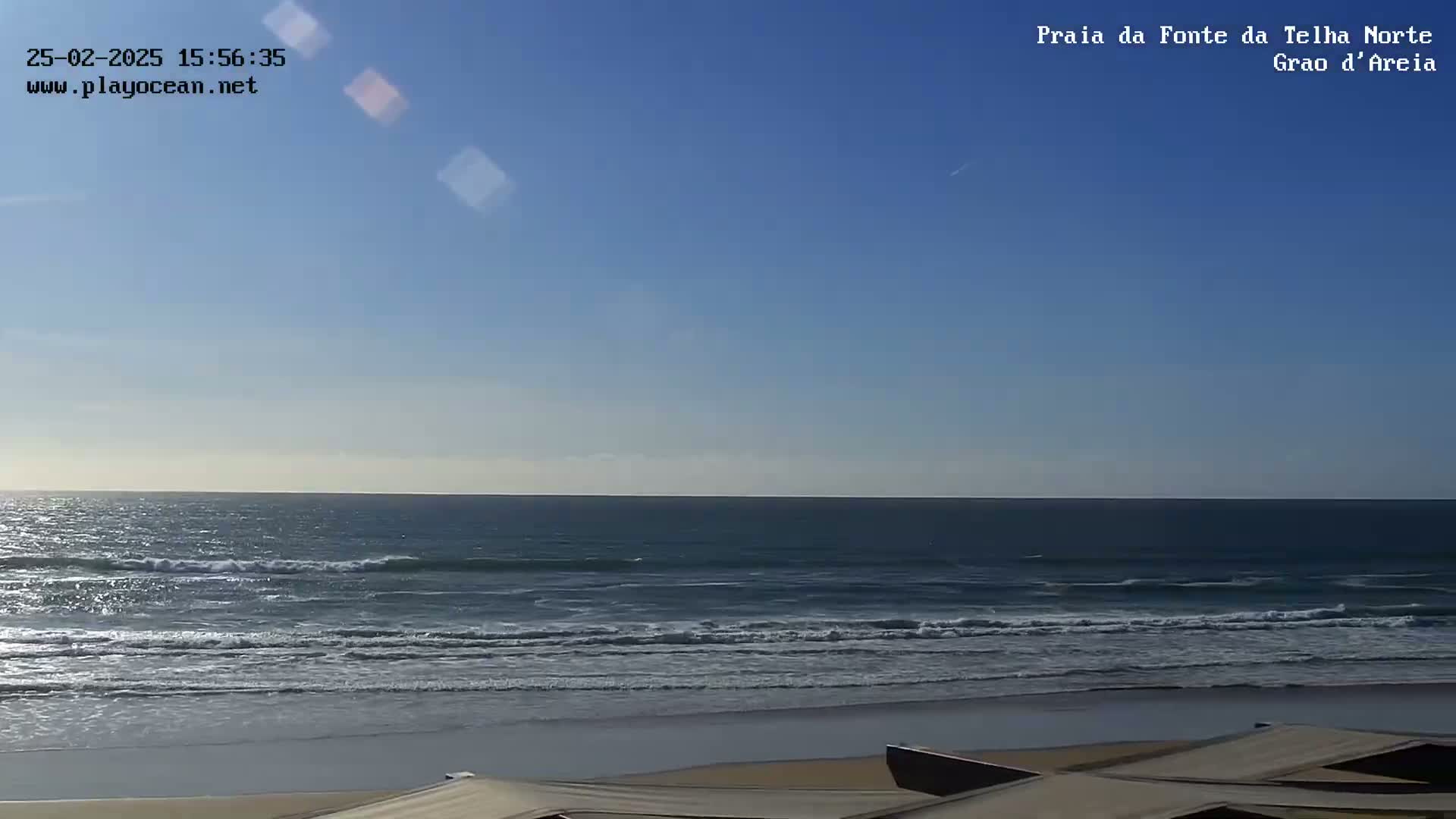 Under a clear blue sky, a calm ocean with small waves laps a sandy beach where several beige beach shelters are partially visible in the foreground.