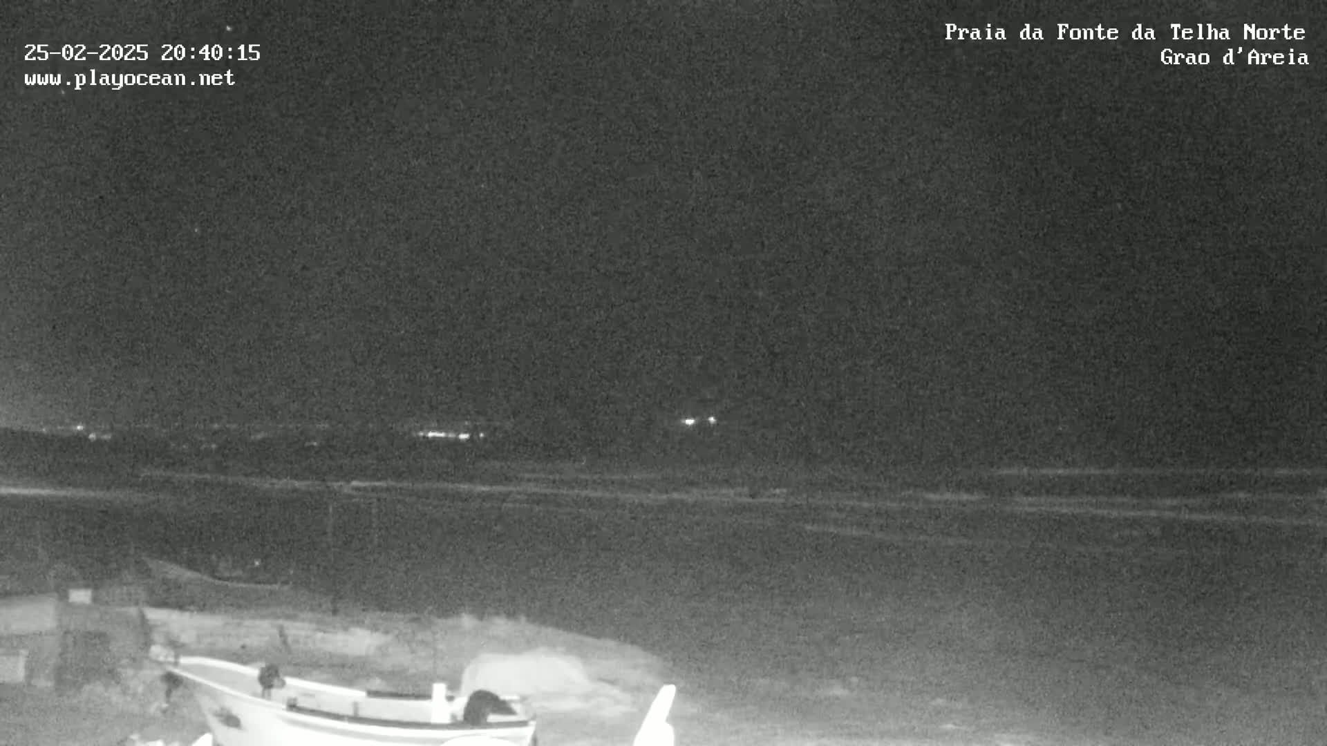 A nighttime, low-light view of a beach shows a dark ocean with a distant shoreline and a boat in the foreground.