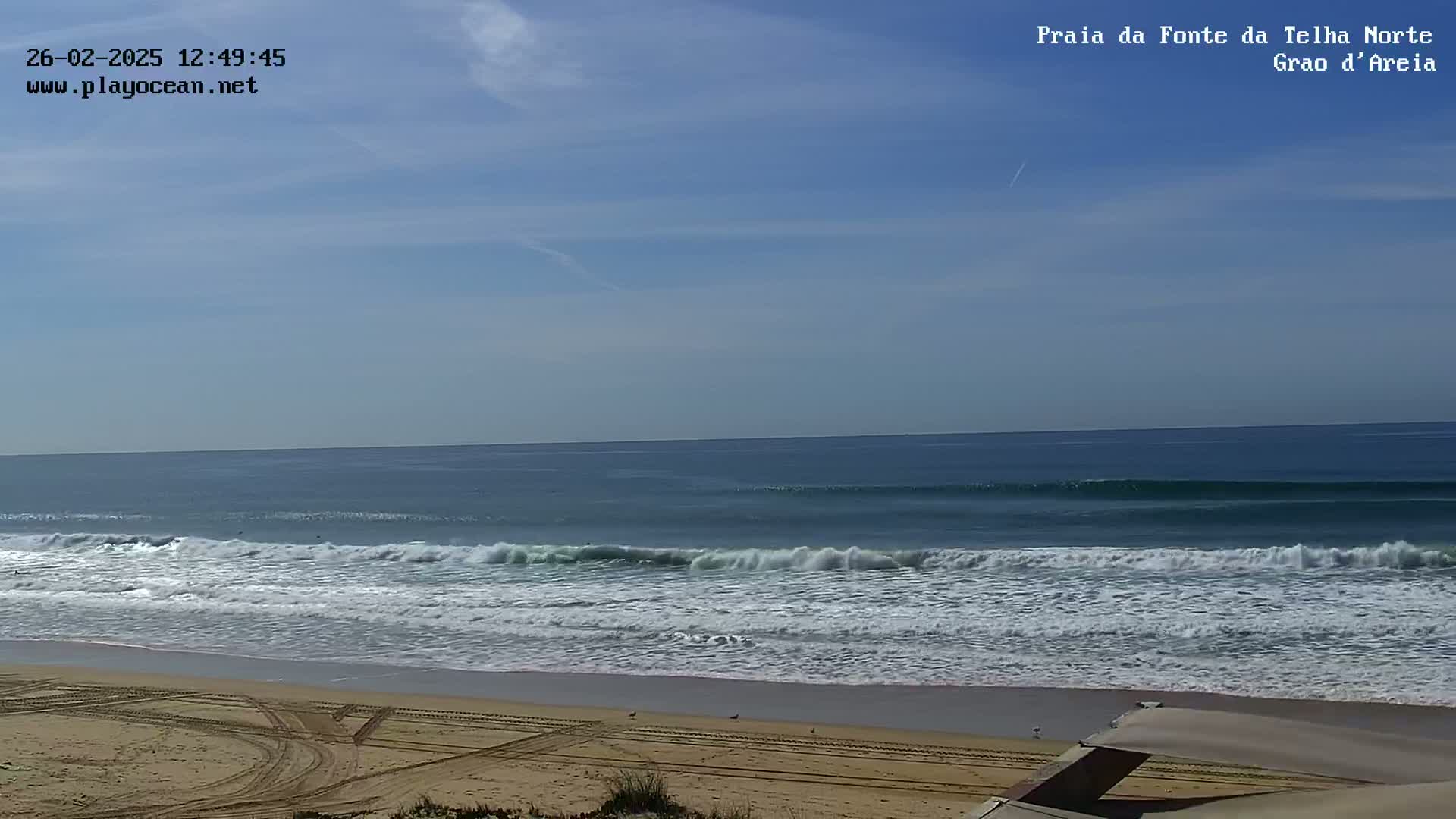 A sandy beach with tire tracks and gentle waves rolling onto the shore under a mostly clear blue sky.