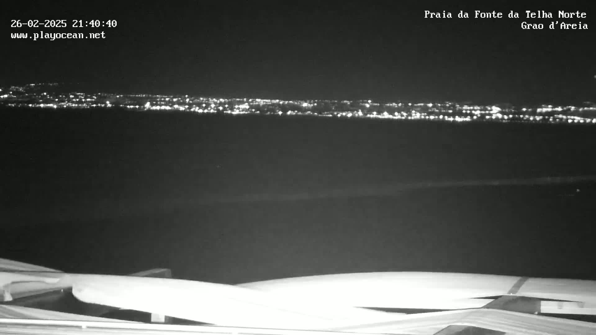 A nighttime long shot of a distant city skyline illuminated by lights, viewed from what appears to be a dark, seaside location.