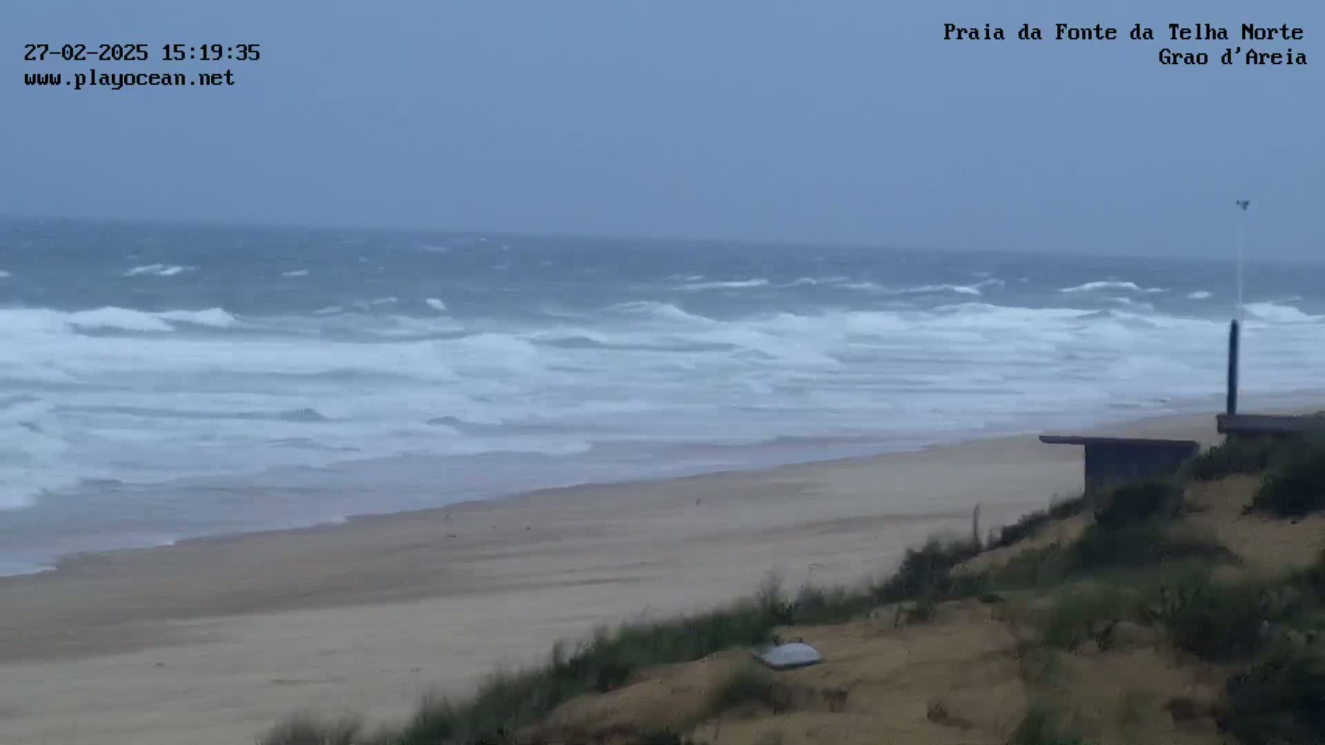 A sandy beach with moderate waves under an overcast sky is viewed from a dune overlooking a structure near the water.