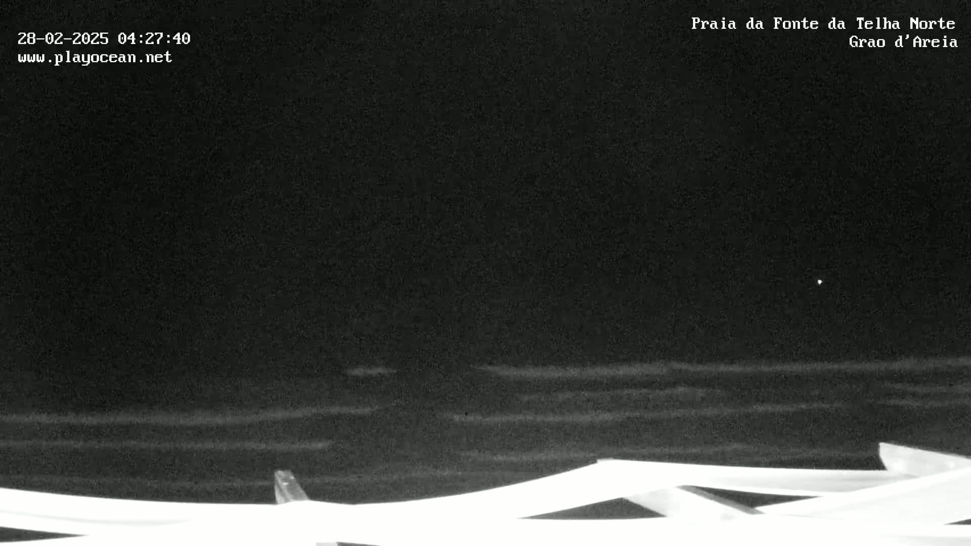 A nighttime view of dark ocean waves gently lapping against a shoreline, partially obscured by a white structure in the foreground.