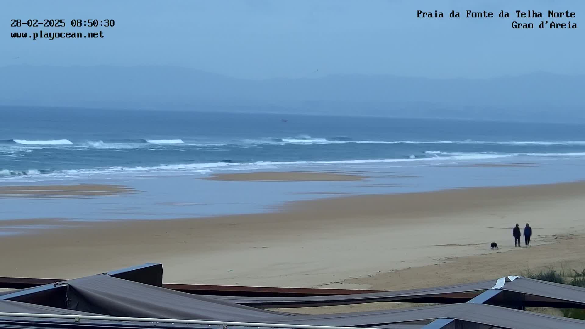 A wide, sandy beach stretches out to a calm ocean with small waves under an overcast sky, where two people walk a dog.