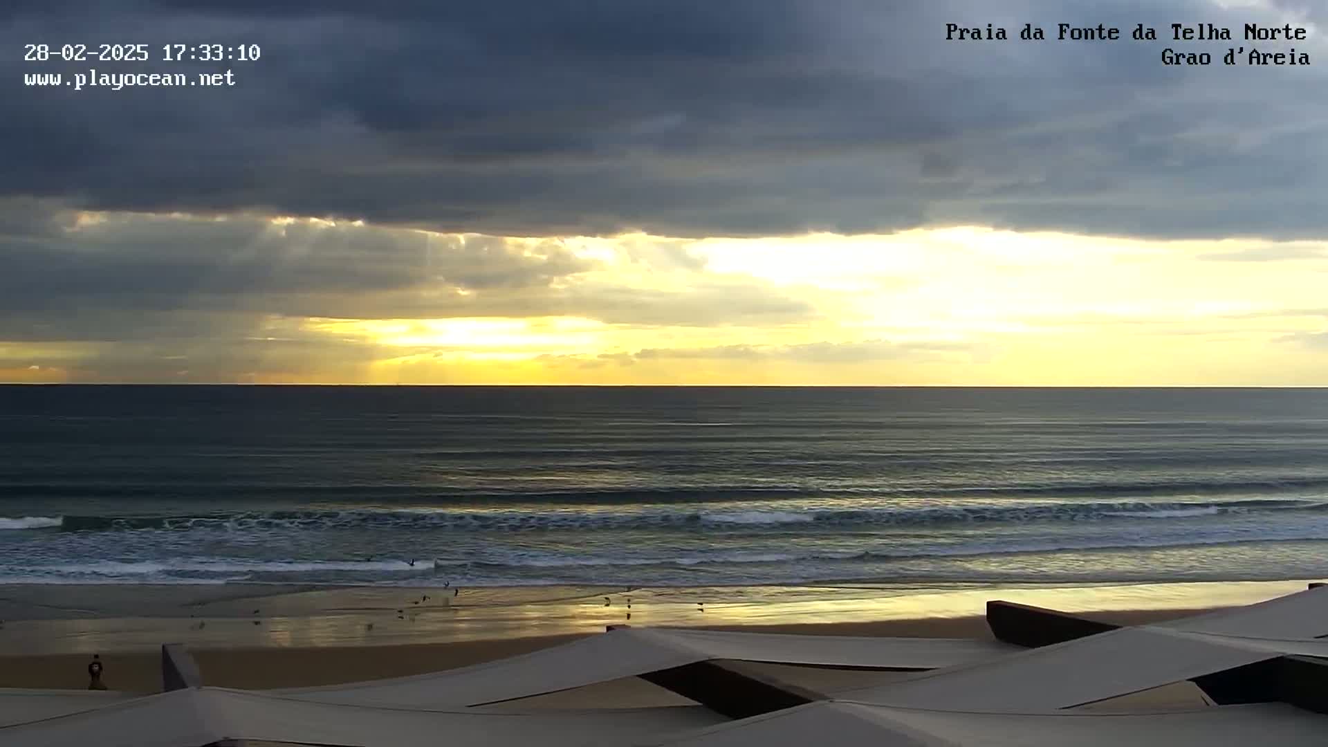 A sunset with sun rays breaking through dark clouds over a calm ocean beach with a sandy shoreline and a few people visible.