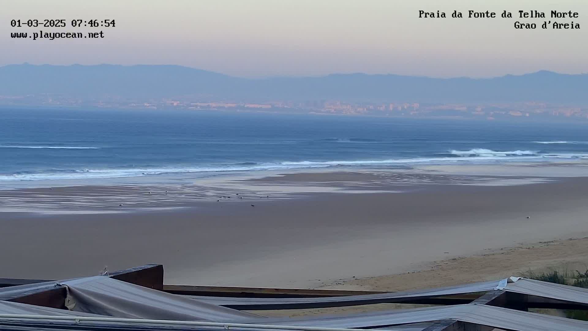 A wide sandy beach stretches out to a calm ocean under a hazy sky with distant mountains visible on the horizon.