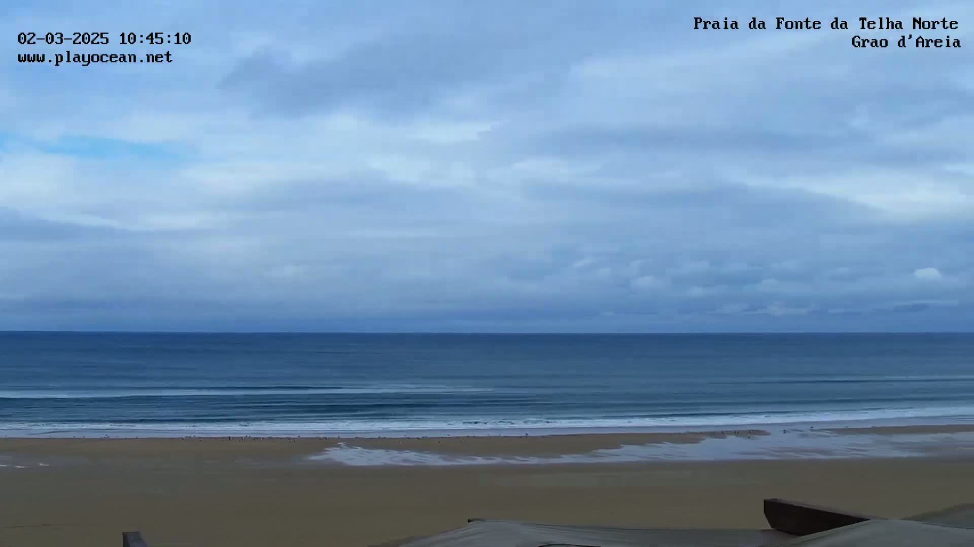 A wide shot shows a sandy beach and a calm ocean under a mostly cloudy sky.