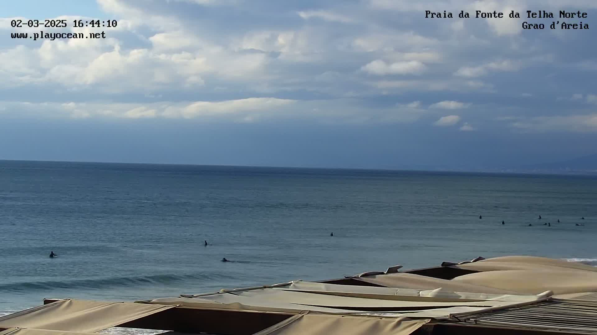 A partly cloudy day shows several surfers in the ocean viewed from a beach with tan-colored canopies in the foreground.