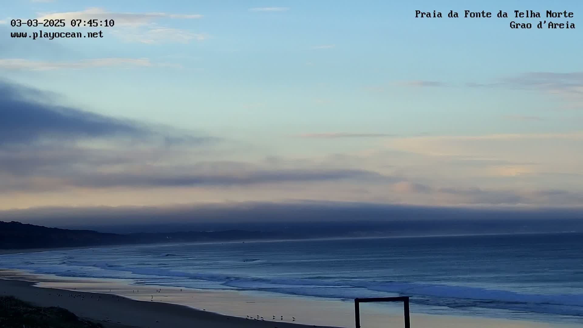 A hazy sunrise illuminates a beach with gentle waves rolling onto the shore under a mostly cloudy sky.
