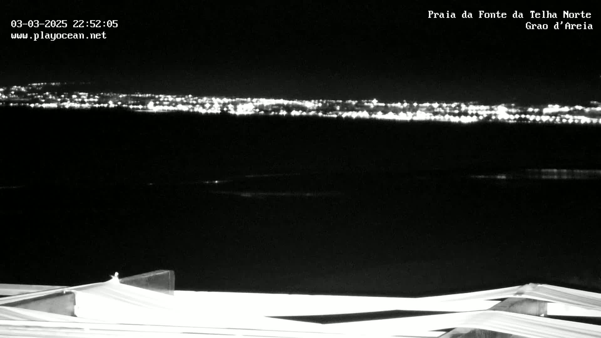 A nighttime long shot shows a distant city skyline illuminated by lights across a dark body of water, viewed from a vantage point with a white structure in the foreground.