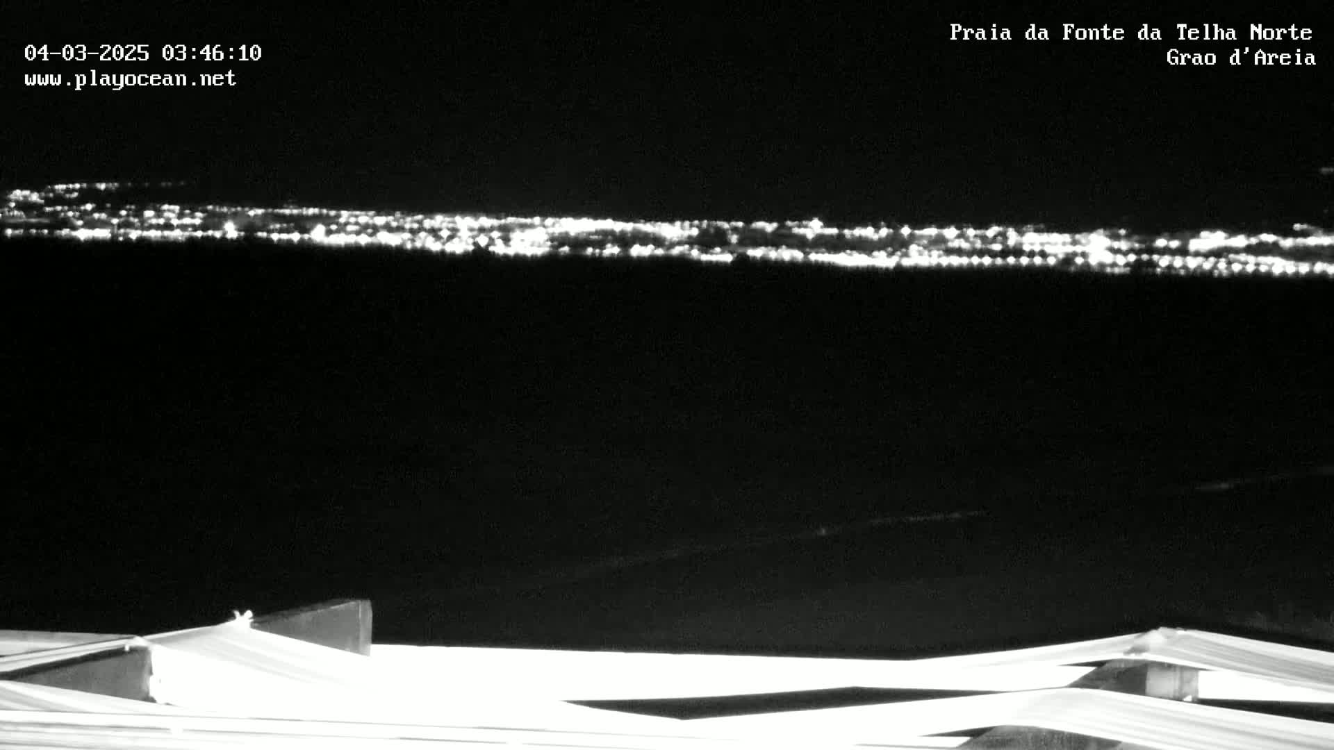 A nighttime long shot shows a distant city skyline illuminated by lights, viewed from a vantage point with a white structure in the foreground under a clear, dark sky.