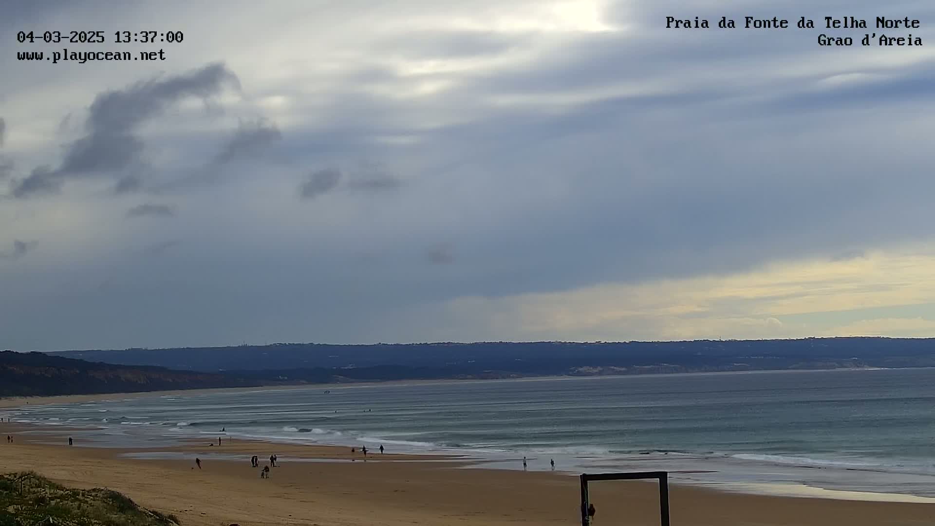 A mostly cloudy sky overlooks a long, sandy beach with gentle waves and a few people scattered along the shore.