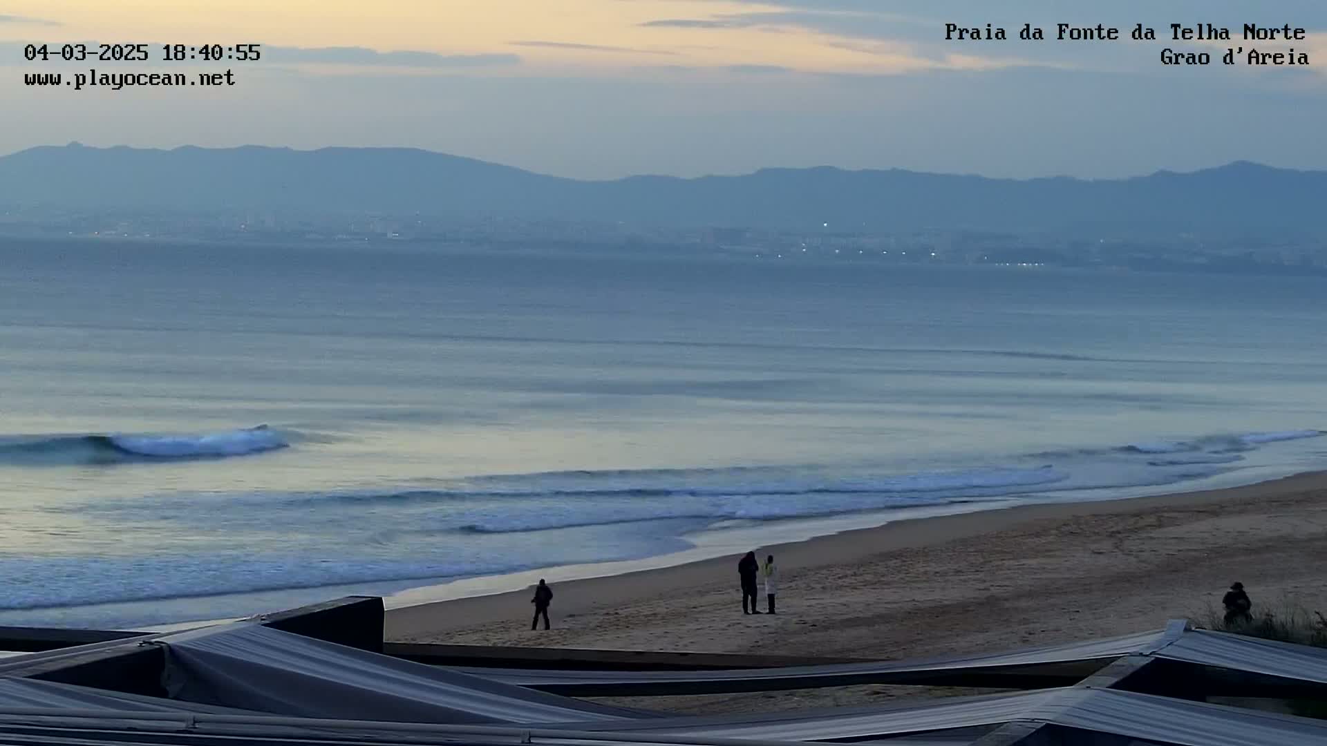 A sandy beach with gentle waves under a twilight sky, a few people are standing on the shore, and distant mountains are visible in the background.
