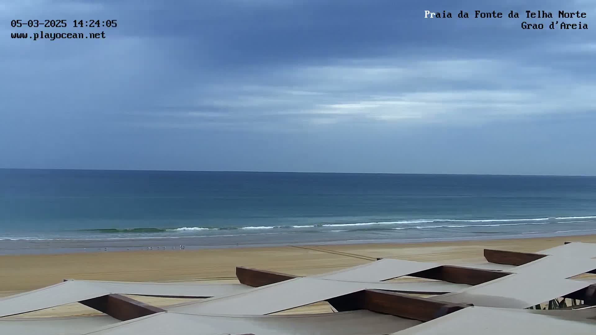 A sandy beach with calm ocean waves under a mostly cloudy sky is viewed from above a series of shaded outdoor lounge structures.