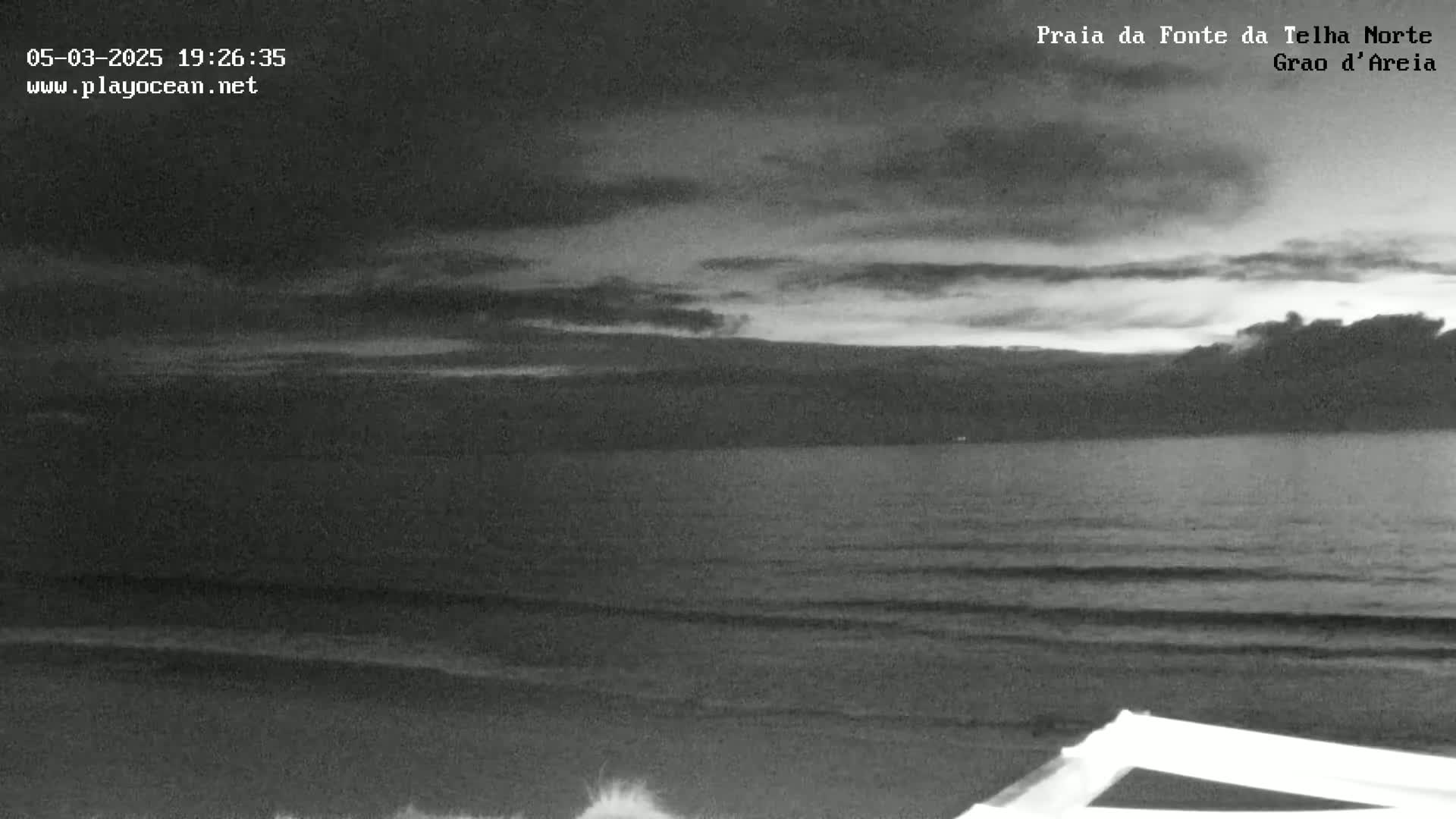 A grayscale image shows a dark, overcast sky over a calm ocean with small waves lapping the shore.
