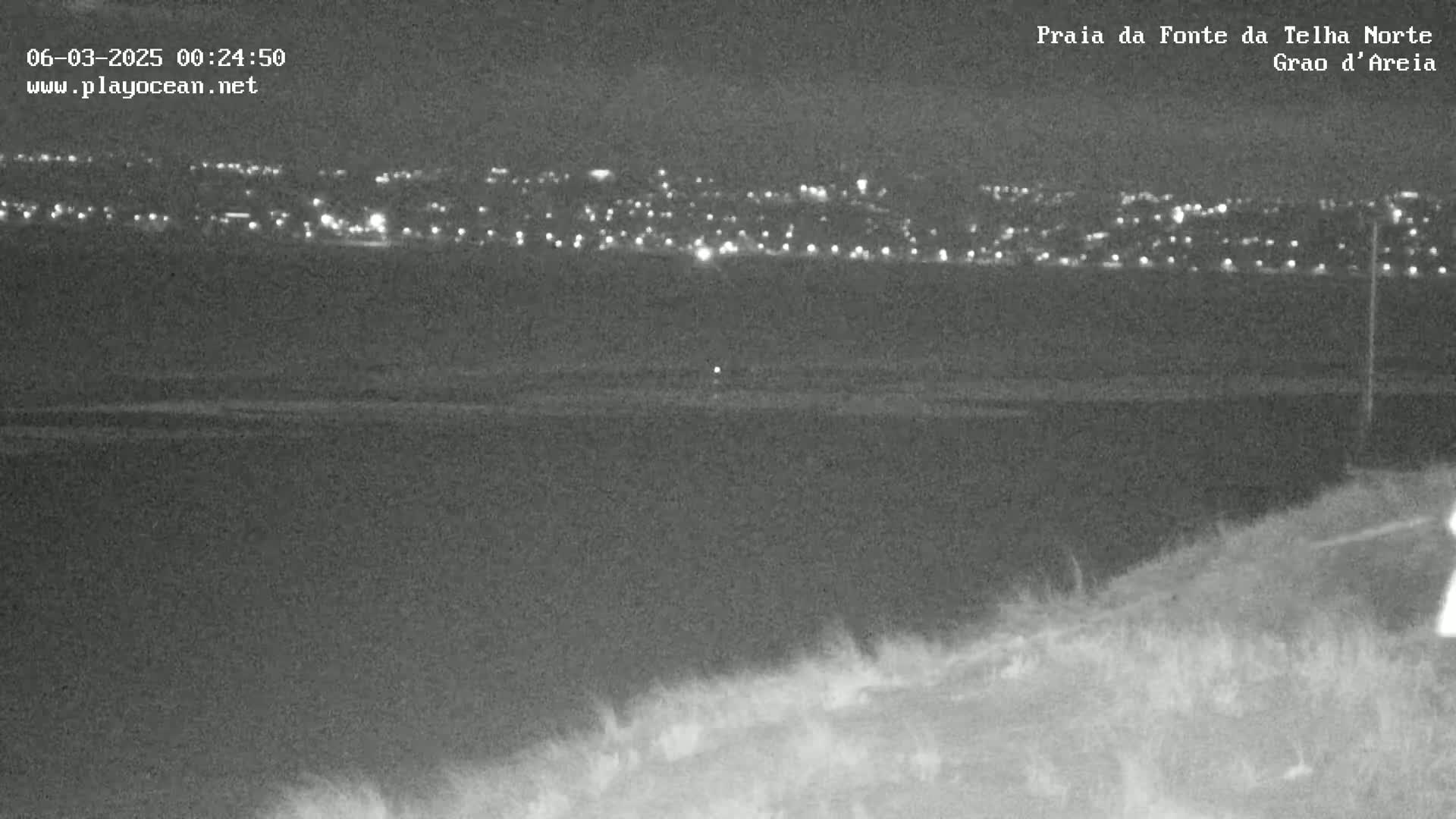 A nighttime, low-light image shows a distant town illuminated with lights across a dark body of water, viewed from a high vantage point on land.
