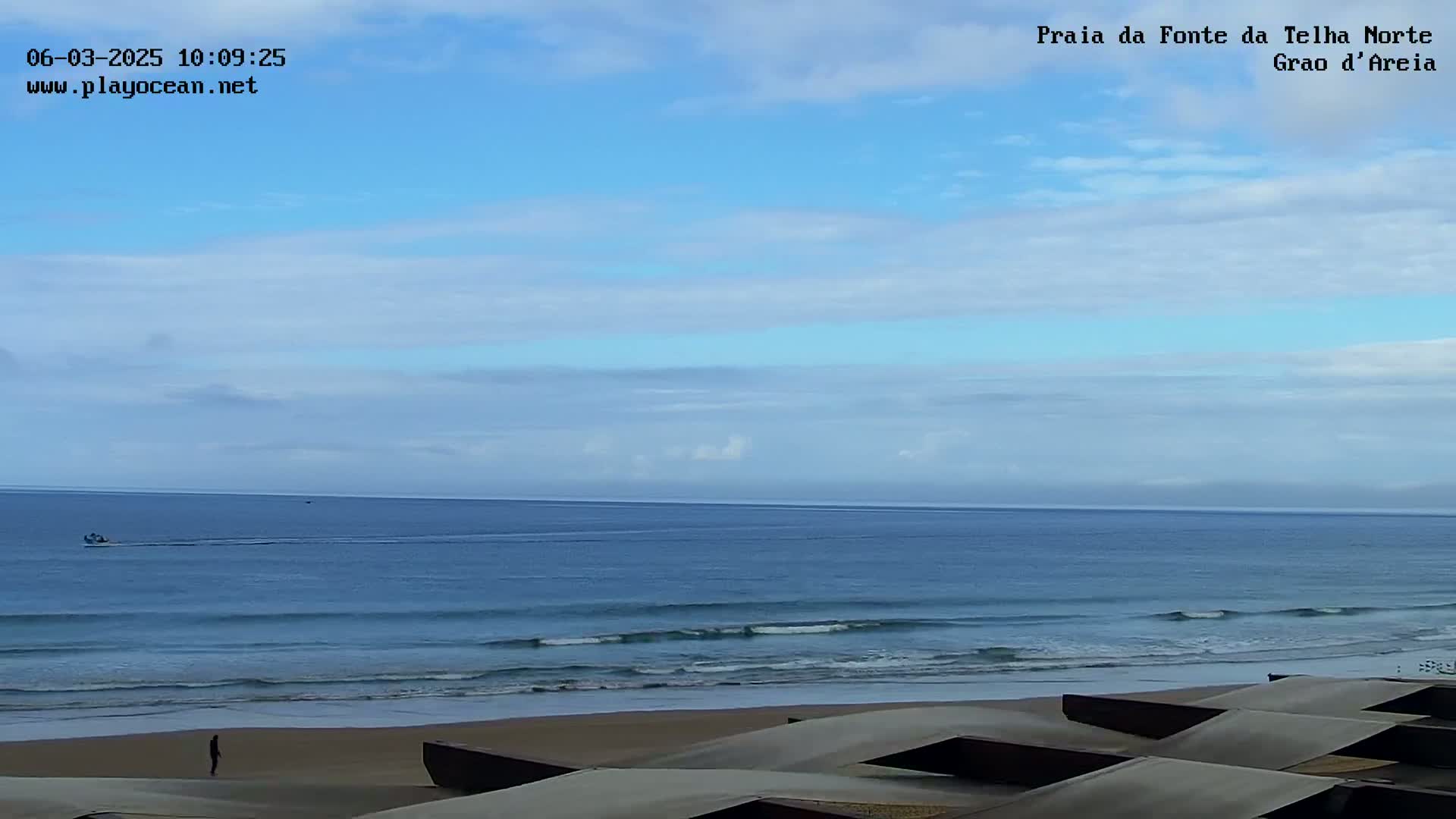 A mostly sunny day at a beach shows a calm ocean with small waves lapping the shore, several dark-colored structures on the sand, and a lone person walking.