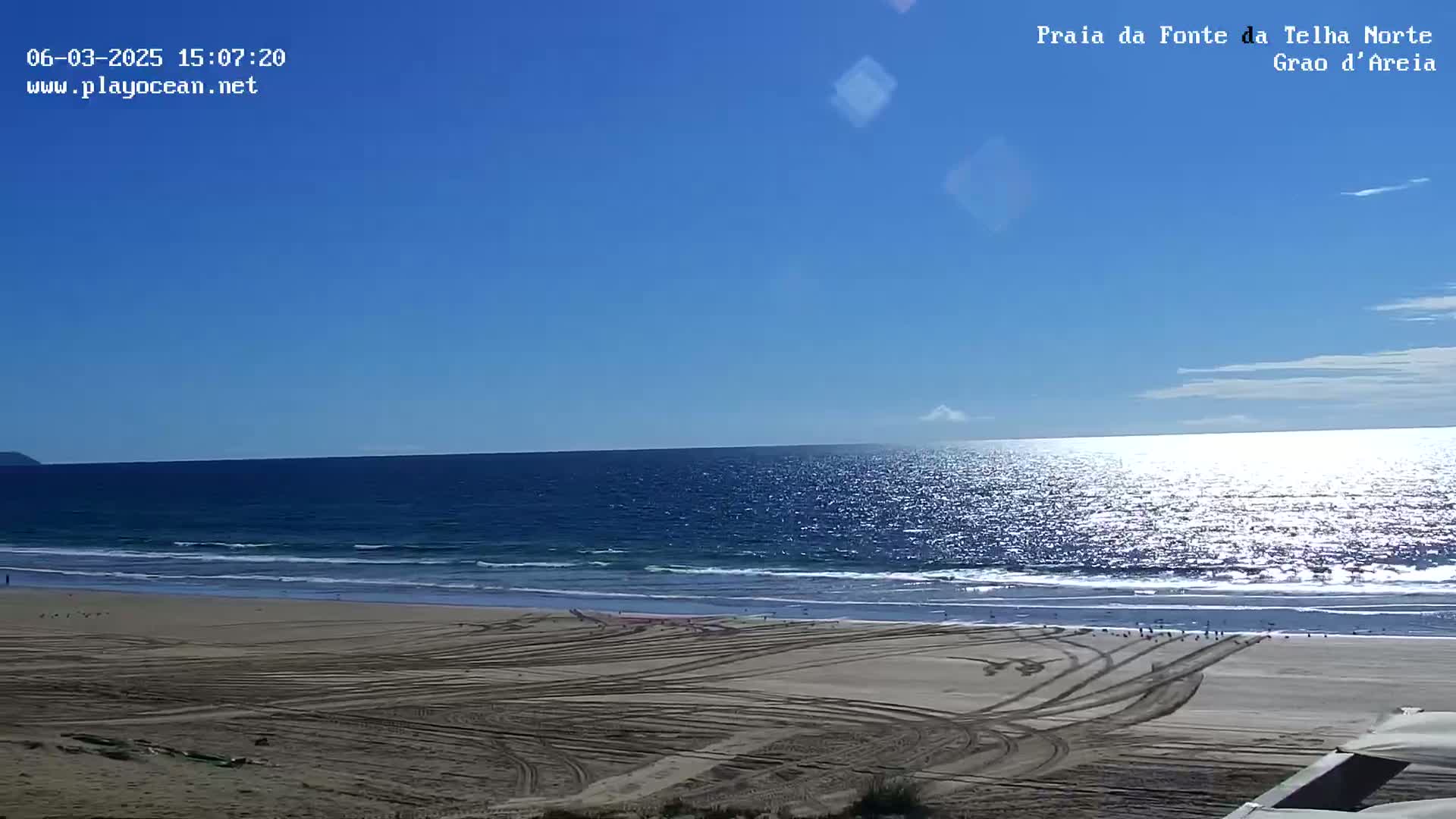 A sandy beach with tire tracks shows a calm ocean under a clear blue sky.