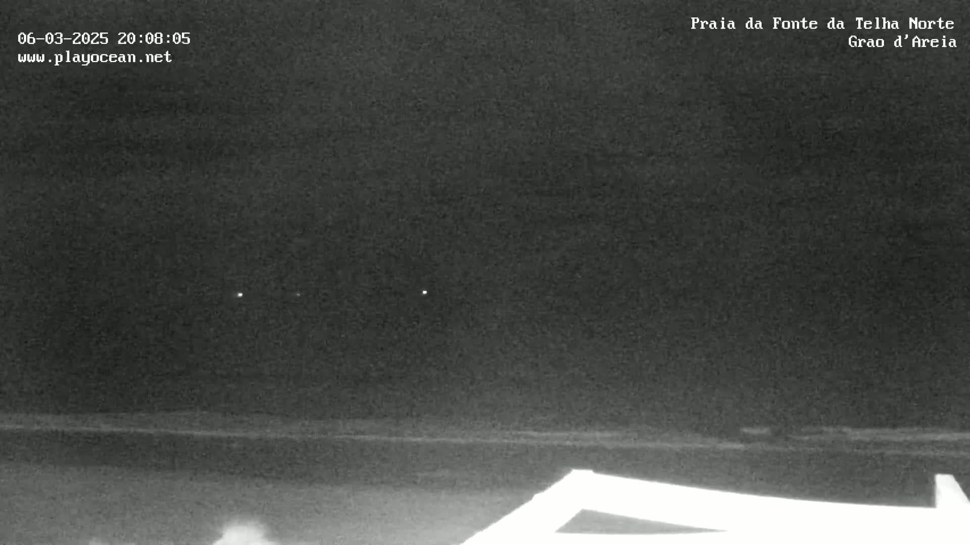 A nighttime view of a dark beach under a cloudy sky with a few distant lights visible.