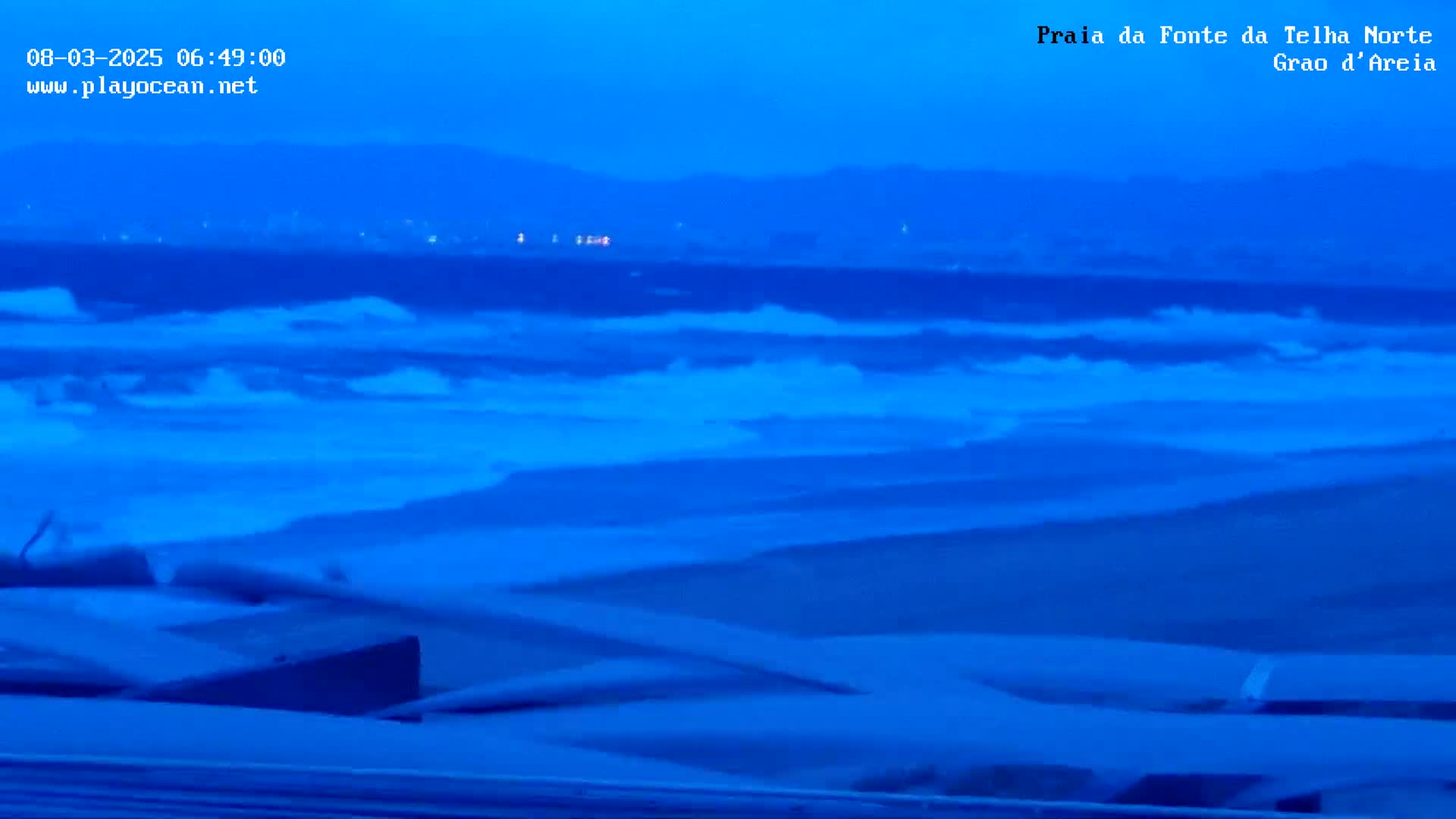 Under a dark blue, pre-dawn sky, waves crash on a beach in front of a distant, dark shoreline.
