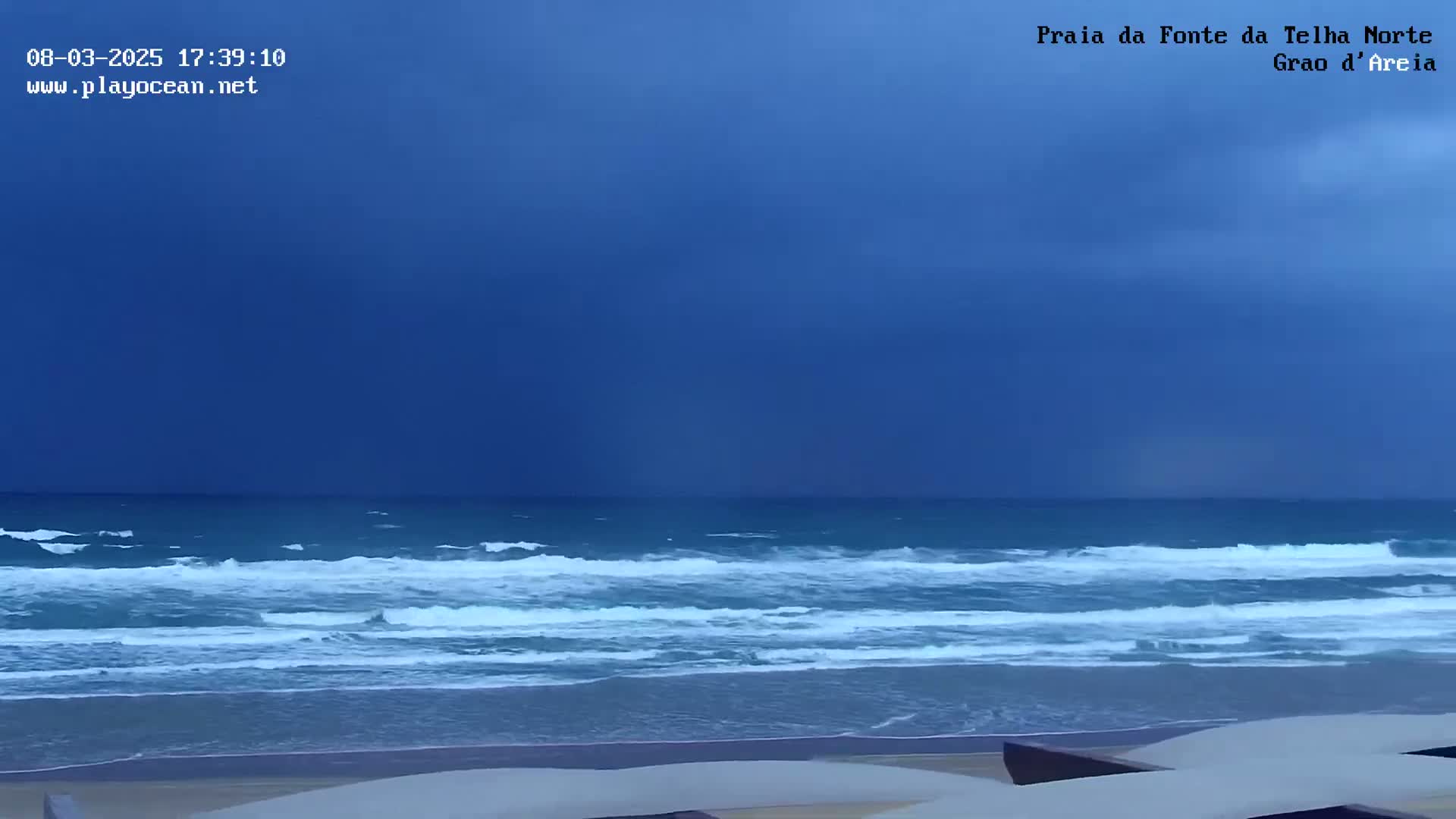 A beach scene under a dark blue, overcast sky shows waves rolling onto the shore.
