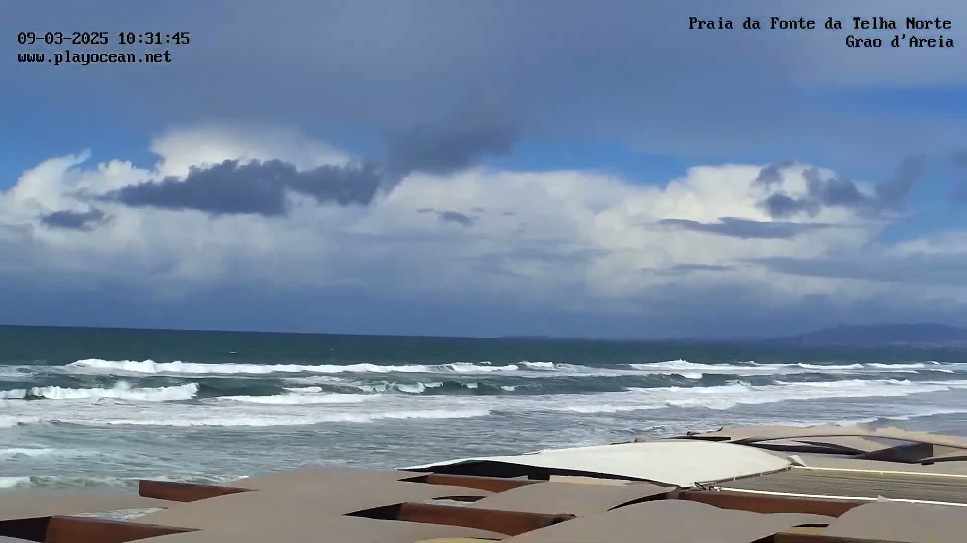 A partly cloudy sky overlooks a beach with moderate waves, and several tan-colored structures with white tops are visible in the foreground.