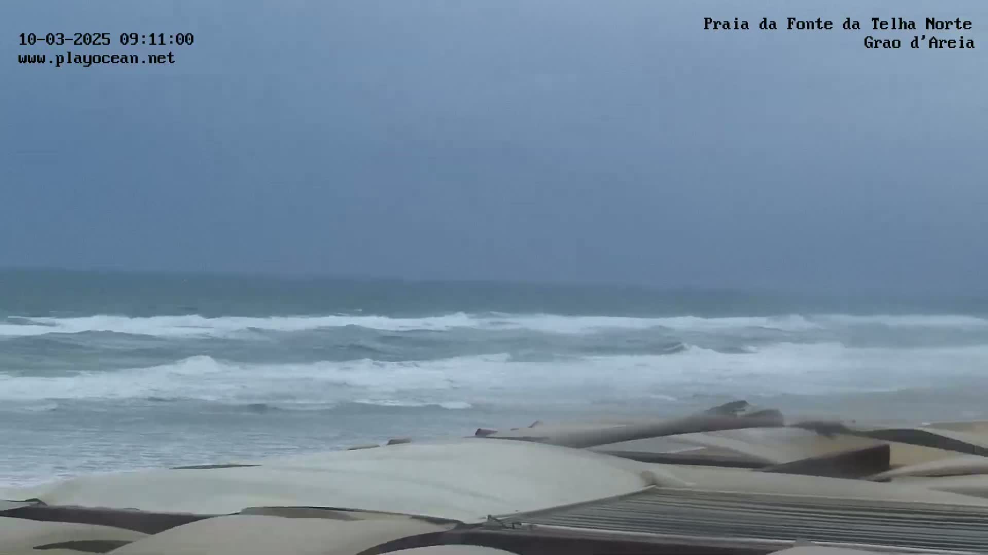 A stormy ocean with choppy waves crashes onto a beach covered with what appears to be sand-colored structures.