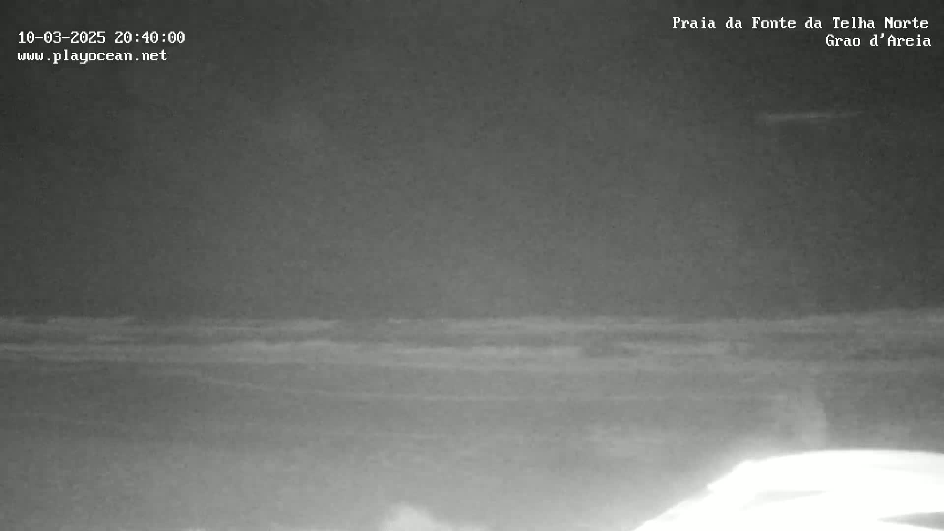 A nighttime, low-light image shows a dark beach with gently moving waves under a cloudy sky.