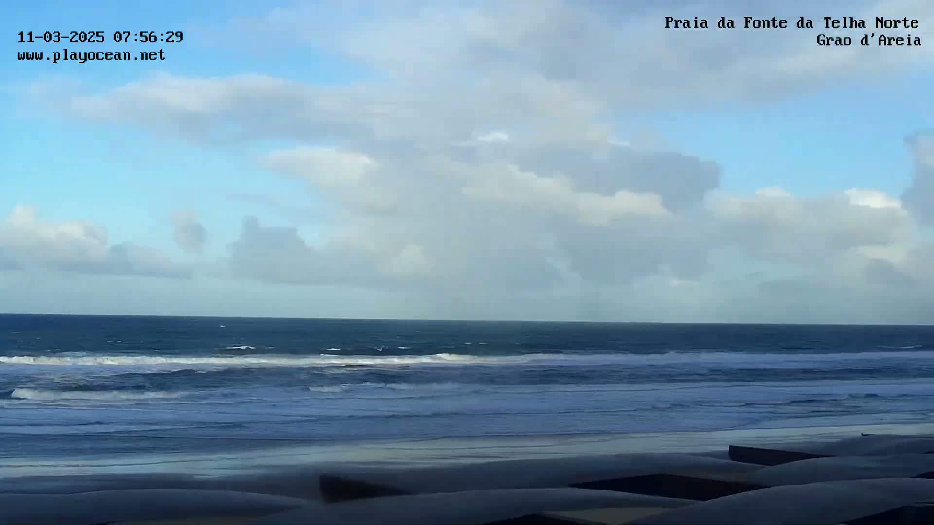 A partly cloudy sky overlooks a dark blue ocean with small waves breaking on a sandy beach.