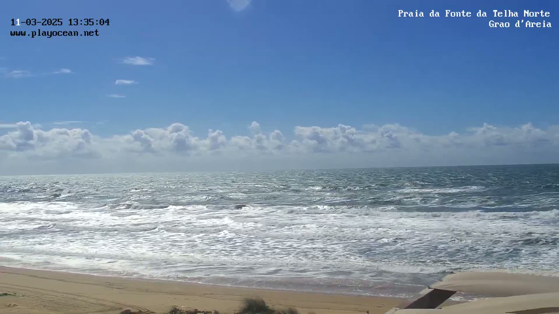 A sandy beach meets a moderately wavy ocean under a mostly sunny sky with scattered cumulus clouds.