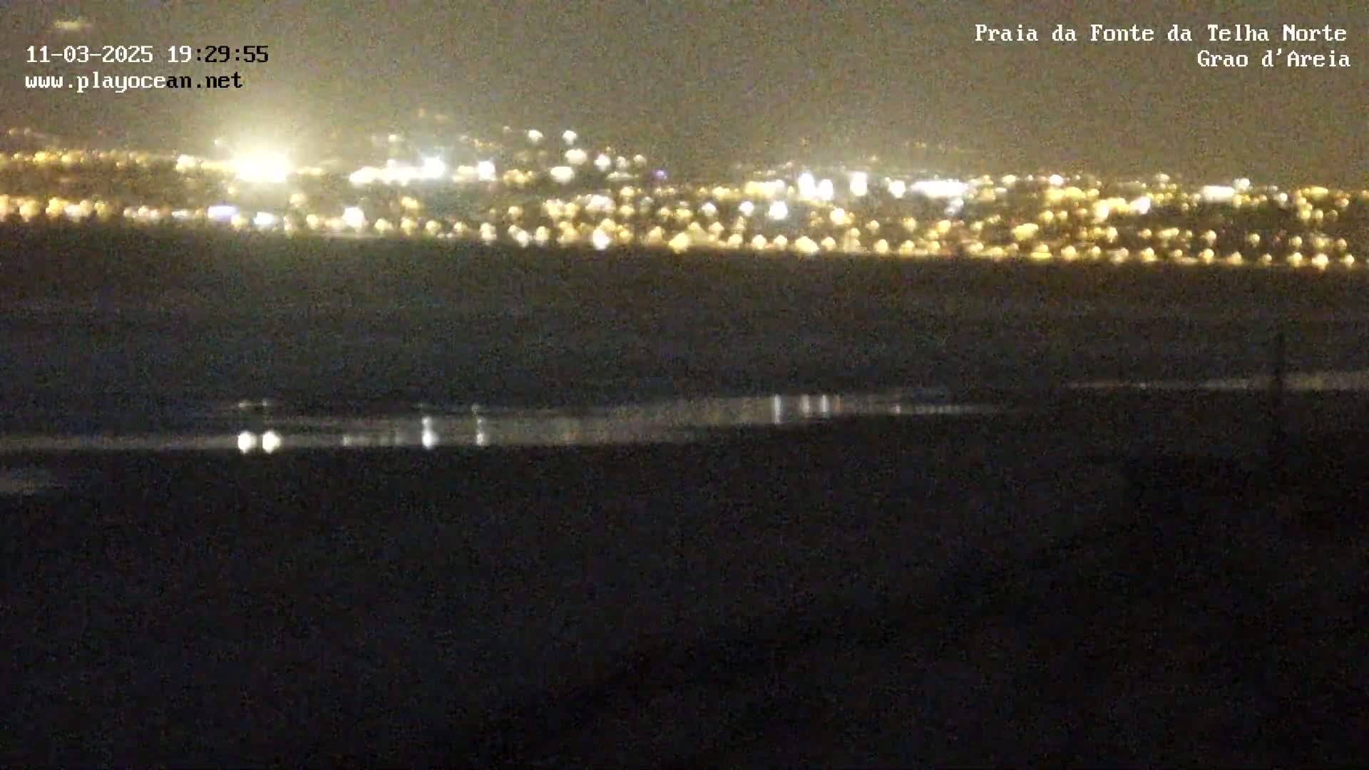 A nighttime view shows a distant city skyline illuminated with lights across a dark, calm body of water.