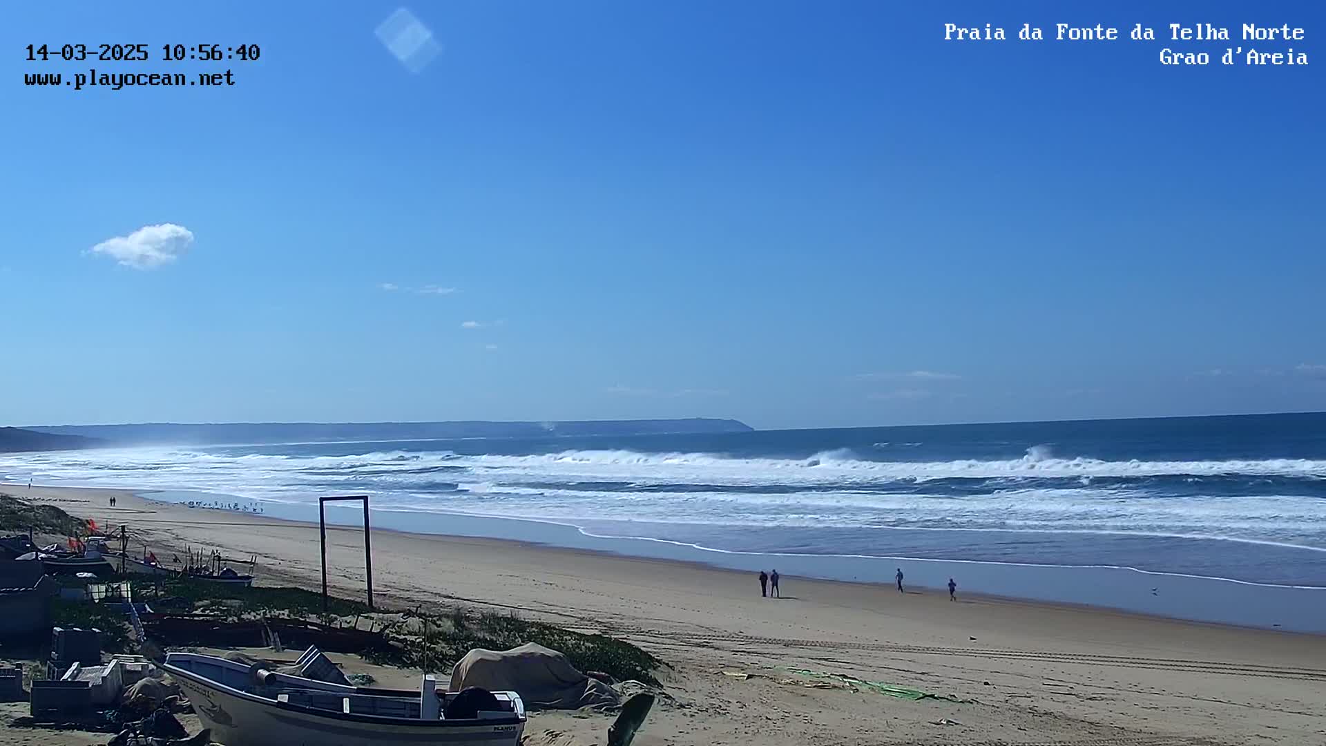A sandy beach with several people walking along the shoreline, boats in the foreground, and waves breaking in the ocean under a clear blue sky with a few small clouds.