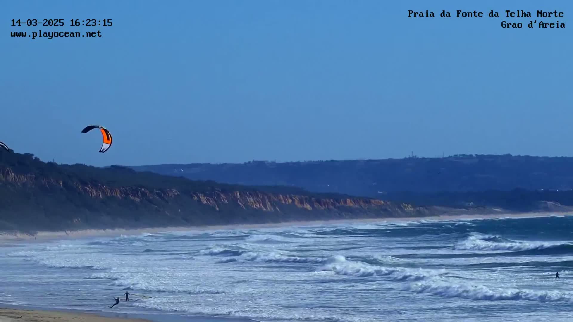 A kite surfer is visible on a beach with moderate waves under a clear blue sky.