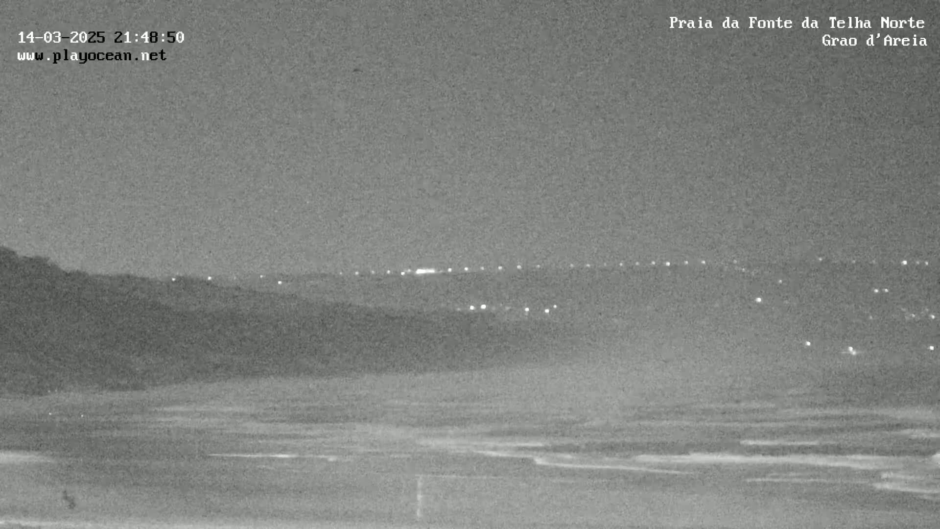 A nighttime, low-light view of a dark beach with gentle waves and a distant line of lights on a hill.