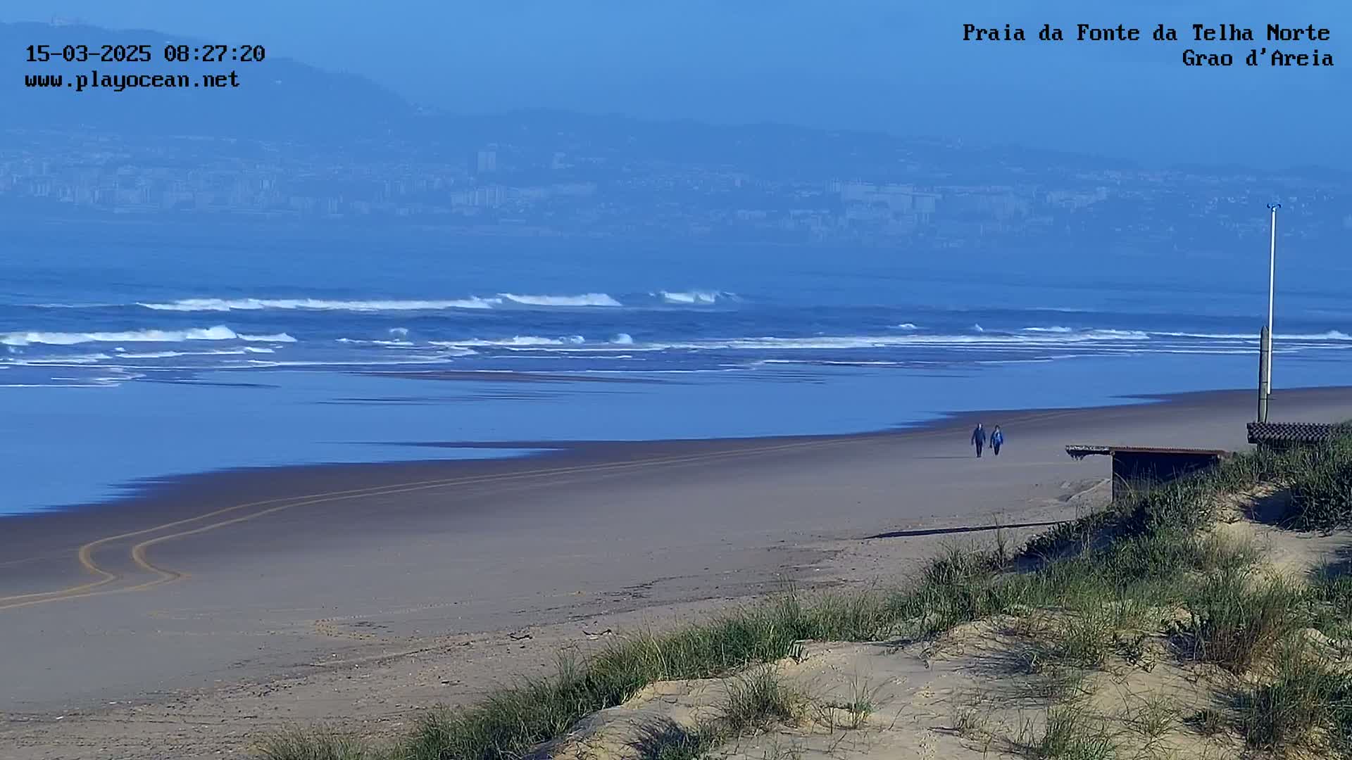 Two people walk along a sandy beach with gentle waves breaking in the ocean under a clear, hazy blue sky.